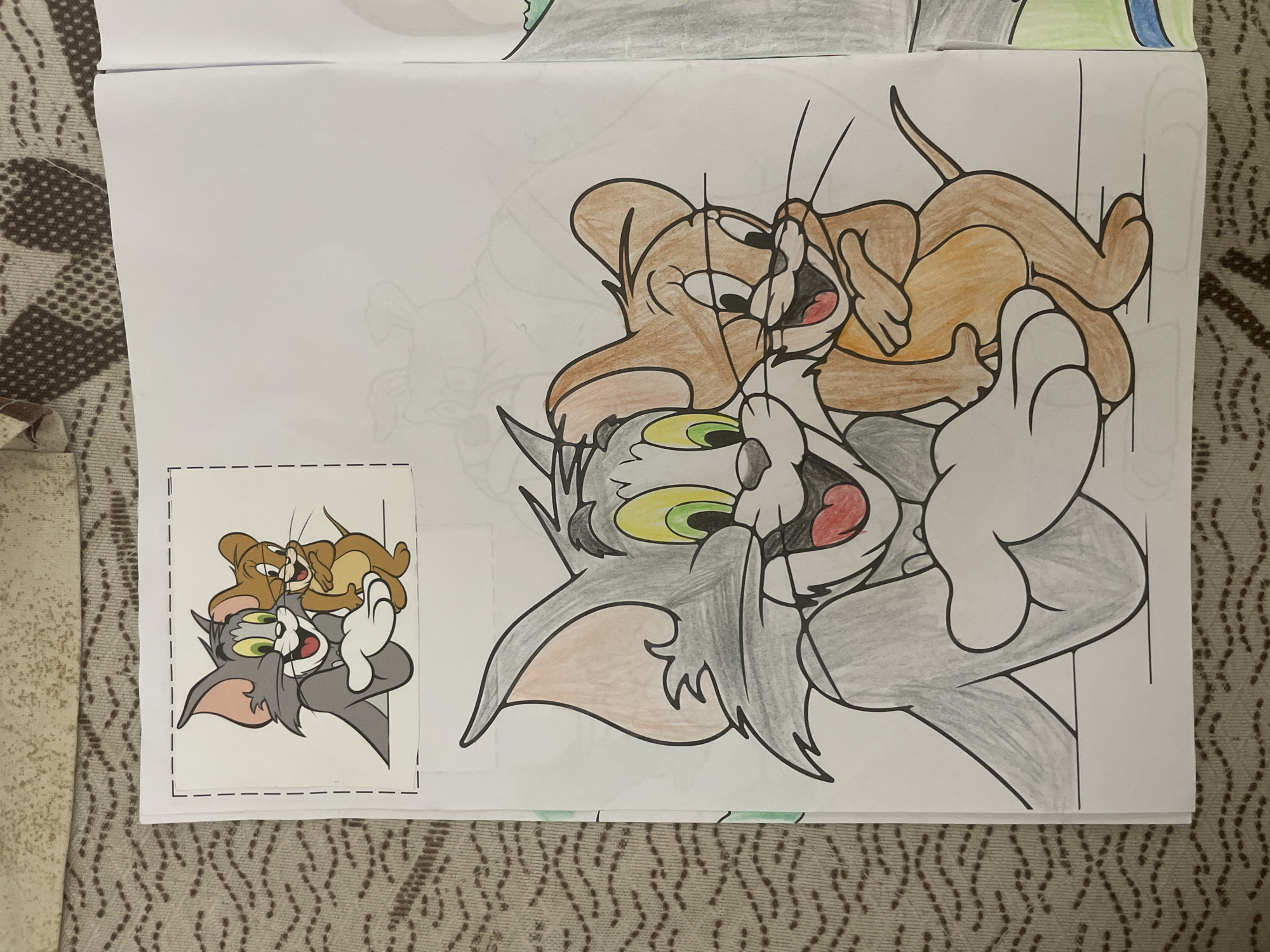 The image displays a coloring book page featuring a large illustration of the iconic cartoon characters, Tom and Jerry, partially colored. Tom, the gray cat, is depicted on his back with a surprised or wide-eyed expression, while Jerry, the brown mouse, is playfully positioned on top of him, also appearing excited or mischievous. Tom's fur is colored in shades of gray, his eyes are green and yellow, and his mouth is open with a red interior. Jerry's body is filled with brown and light brown tones. Both characters have black outlines, and their limbs are intertwined, suggesting a dynamic moment of their classic chase or interaction.
Adjacent to the main illustration, on the lower left of the page, is a smaller, fully colored reference image of the exact same scene. This reference image is surrounded by dashed lines, indicating it was intended to be cut out. The presence of both the partially colored large drawing and the fully colored reference image suggests an ongoing coloring activity.
The coloring book itself rests on a patterned, fabric surface, likely a rug or blanket, which provides a domestic setting for the activity. No other discernible text or specific location cues beyond the home environment are visible. The colors used are vibrant and consistent with the traditional appearance of the characters.