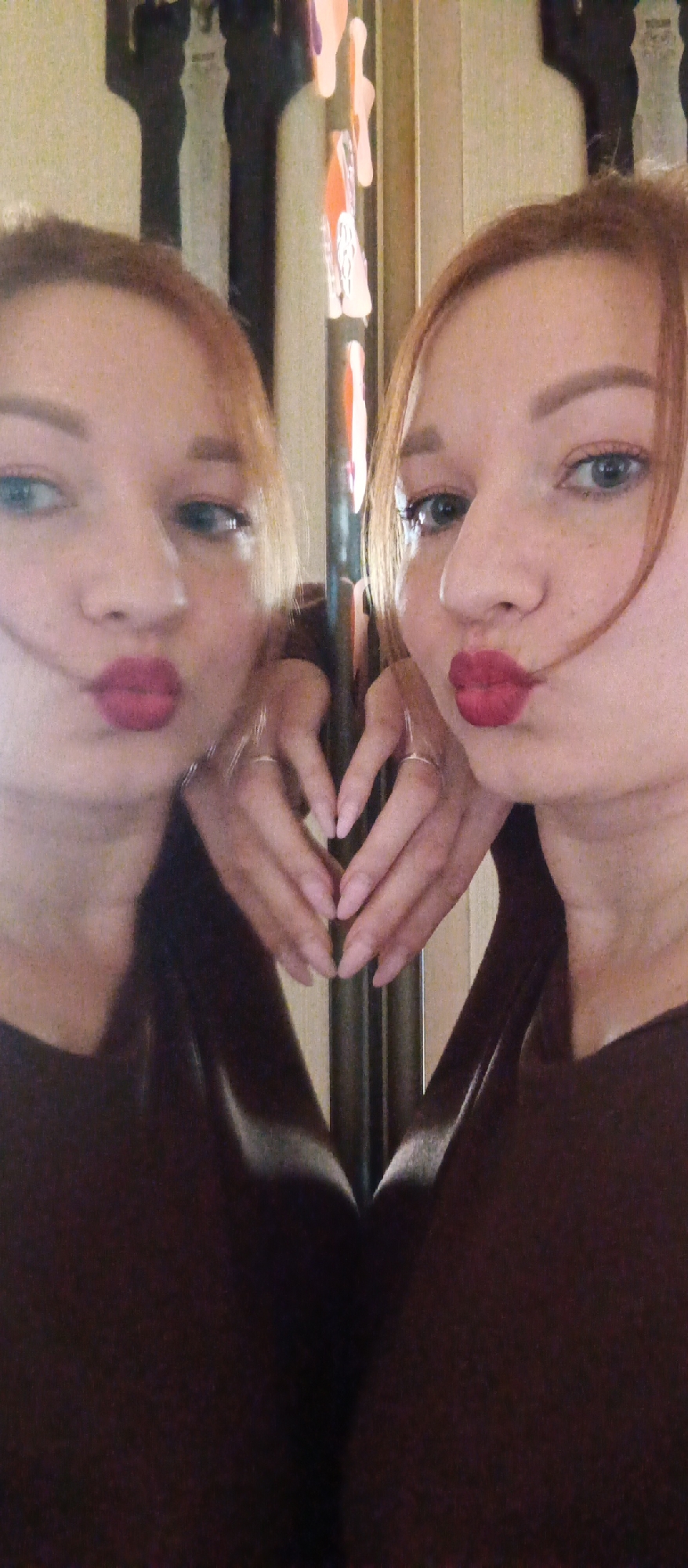 An indoor, eye-level mirror selfie features a woman with light reddish-brown hair and blue eyes, captured alongside her reflection. The image is split vertically by a dark line, indicating the edge of the mirror.
The woman is on the right side of the frame, facing slightly left and upwards, while her reflection is on the left, facing right. Both her real image and reflection are making a "kiss" face with bright red, pursed lips. Her blue eyes are visible, and she wears subtle eyeliner and possibly eyeshadow. Freckles are visible on her cheeks in the original image and crops. Her hair is parted and frames her face, with some strands falling near her cheeks. She is wearing a dark, possibly maroon or dark brown, top or dress.
Her hands, with light pink fingernail polish, are brought together in the center of the frame, forming a heart shape. Rings are visible on the ring fingers of both her real hands and their reflection.
The background behind the woman and her reflection is a light, neutral-toned wall with decorative elements hanging vertically. These decorations appear to be abstract shapes or cutouts in shades of orange, white, and purple, contributing to a slightly playful atmosphere. The lighting suggests daytime or well-lit indoor conditions. There is no visible text in the image.