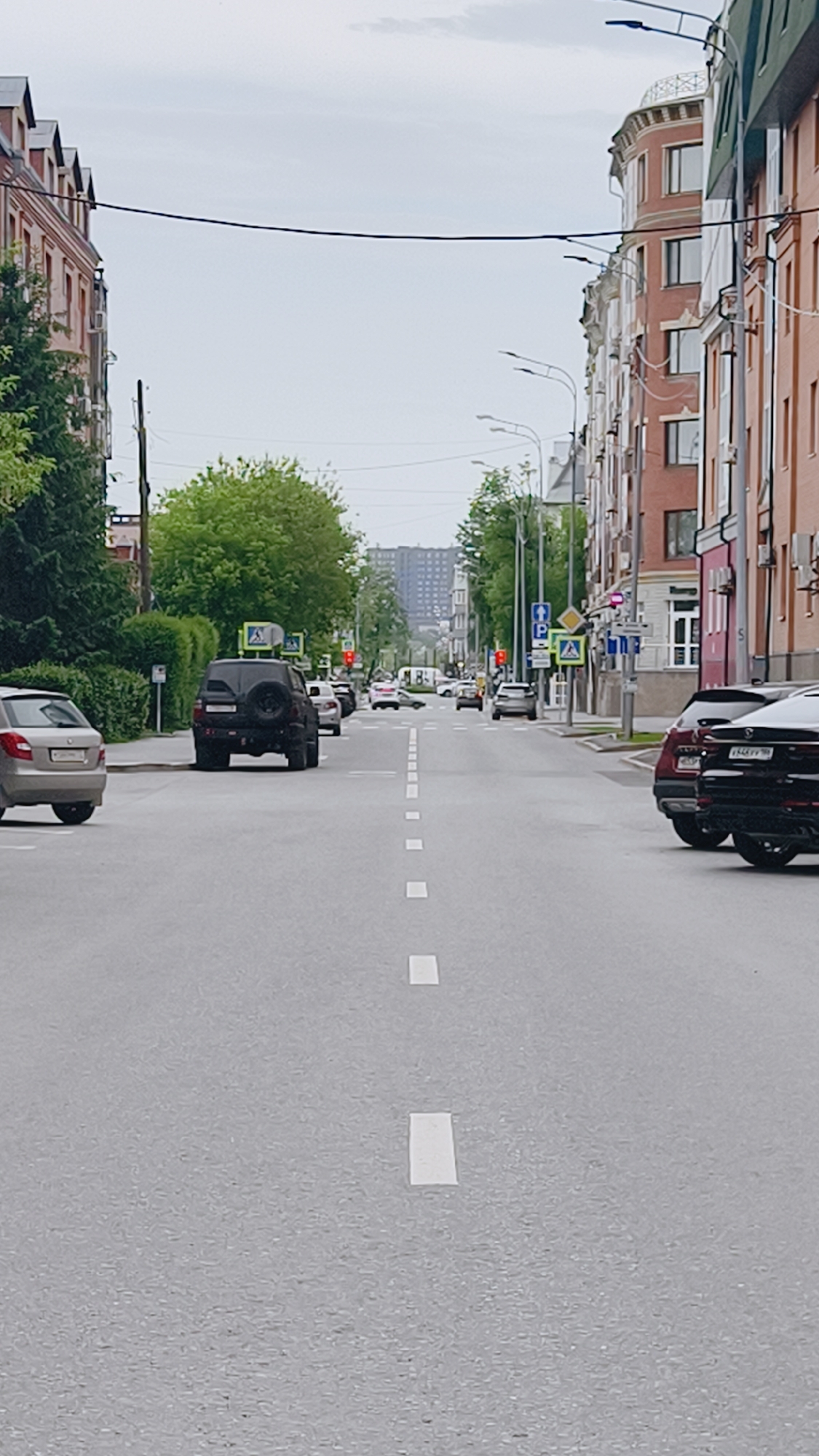 The image depicts a street in Tyumen, Russia. The camera is positioned in the middle of a straight, wide road with white dashed lines marking lanes. Buildings line both sides of the street, constructed of red brick with multiple stories. Trees are interspersed along the sidewalks, adding greenery to the urban scene.
Several cars are visible: one parked on the left with a spare tire mounted on the rear and others are parked on the right side of the road. Traffic lights show a red signal. Pedestrian crossing signs are also visible.
The sky is overcast, casting a diffused light across the scene. In the distance, taller buildings can be seen, suggesting a downtown area. Power lines stretch across the sky above the street.