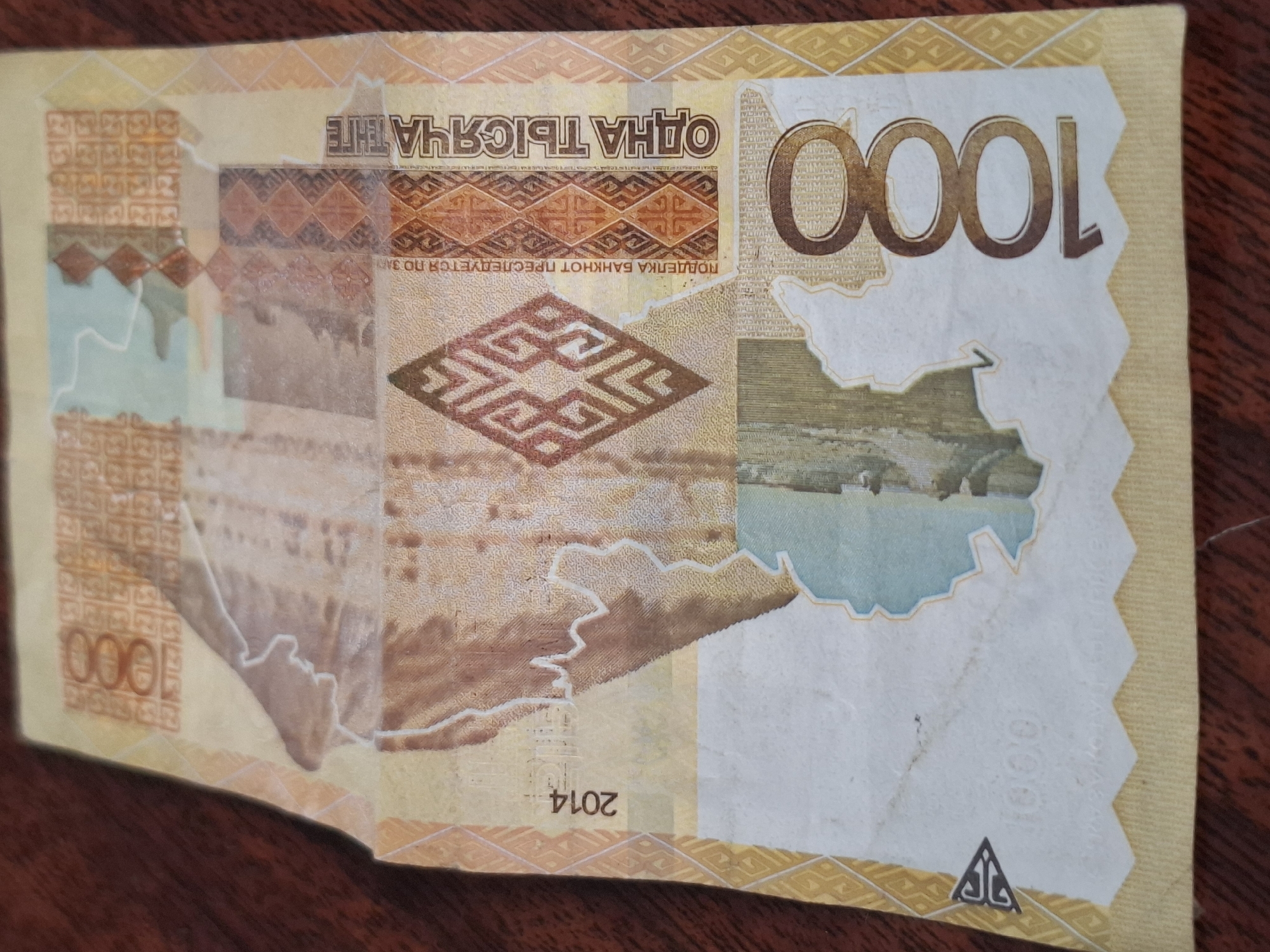 The image displays a Belarusian 1000 Ruble banknote. The banknote features a map of Belarus, a geometric pattern, and the text "АДНА ТЫСЯЧА" which translates to "One Thousand". The year "2014" is also visible. The banknote is lying on a wooden surface, which appears to be a table. There is no indication of the city of Almaty, Kazakhstan, in the image.