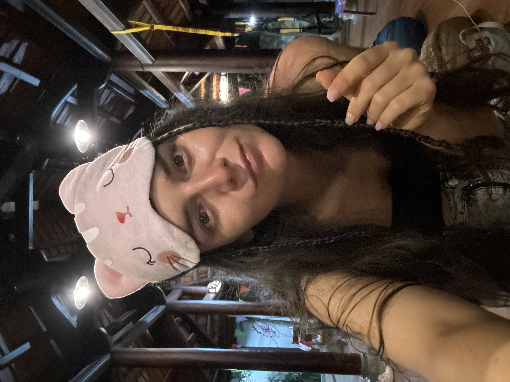 A woman with long dark hair and wearing a pink cat-shaped sleep mask is looking at the camera. She appears to be indoors in a structure with exposed wooden beams. The beams suggest a rustic or traditional architectural style. The picture was taken in Mũi Né, Vietnam.