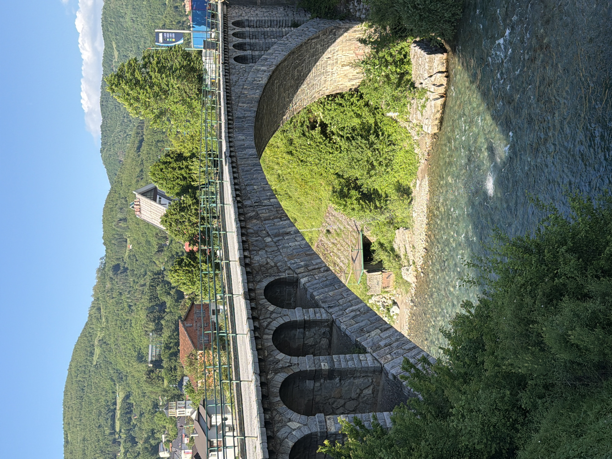 A stone bridge spans a clear, shallow river in Mojkovac, Montenegro. The bridge features arched supports and a green metal railing. Lush green vegetation covers the surrounding slopes and grows alongside the riverbank. Mountains rise in the background under a clear blue sky. Buildings of Mojkovac can be seen nestled among the trees in the distance.