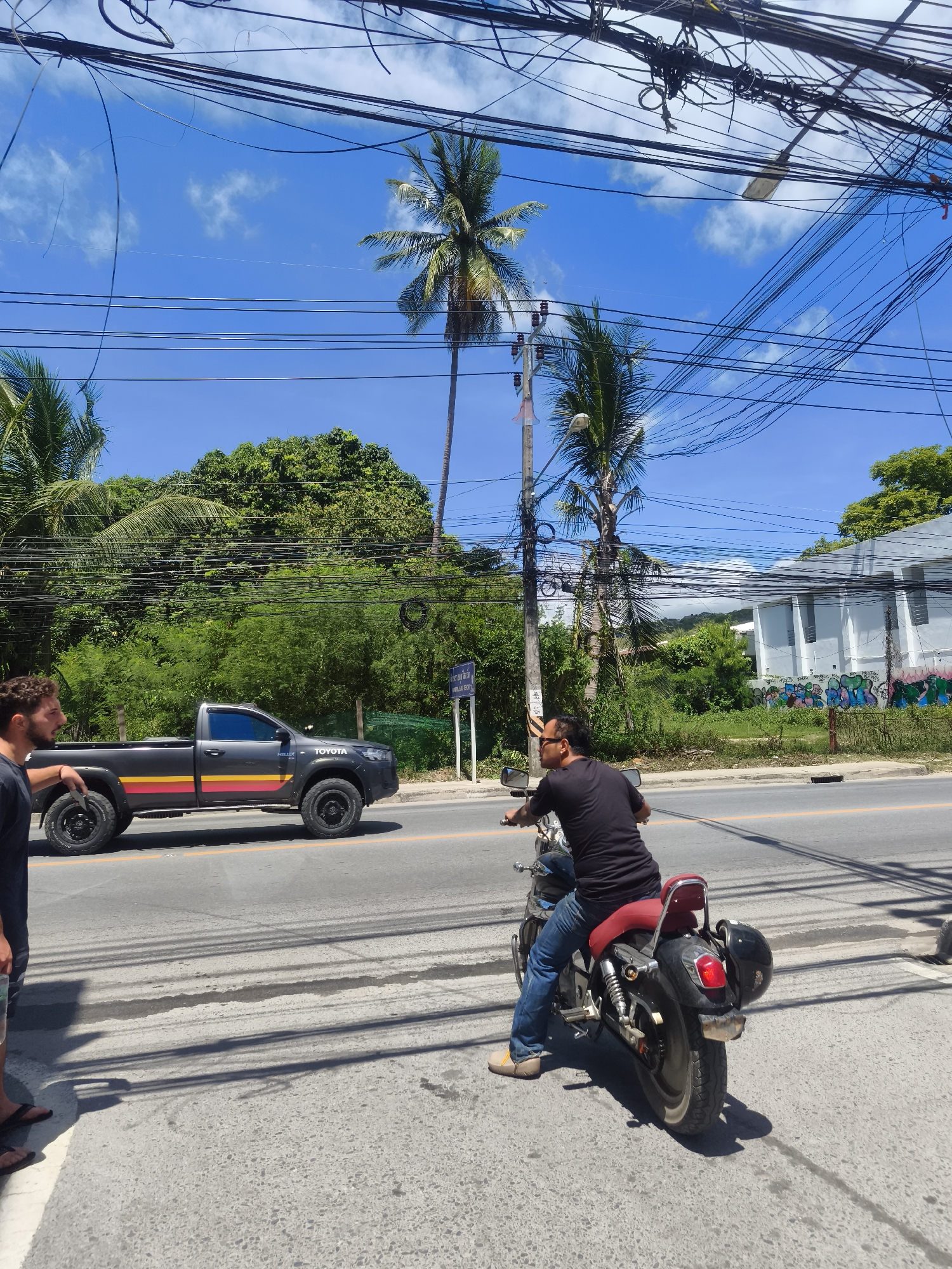 The image shows a street scene in Baan Bo Phut, Thailand. A man stands on a sidewalk on the left side of the frame, facing a gray Toyota Hilux pickup truck with a black roof and distinctive yellow, orange, and red stripes along its side. The truck is stopped in the left lane of the road. A second man is sitting on a motorcycle in the right lane. He is wearing a black t-shirt, blue jeans, and tan shoes. The motorcycle is black with a red seat and has a black helmet strapped to the back. The road is paved and has a yellow dividing line. Across the street from the sidewalk are trees, greenery, and a white building with graffiti on the wall. Palm trees are visible in the background, as are numerous overhead power lines. The sky is blue with some clouds. The lighting suggests it is daytime.