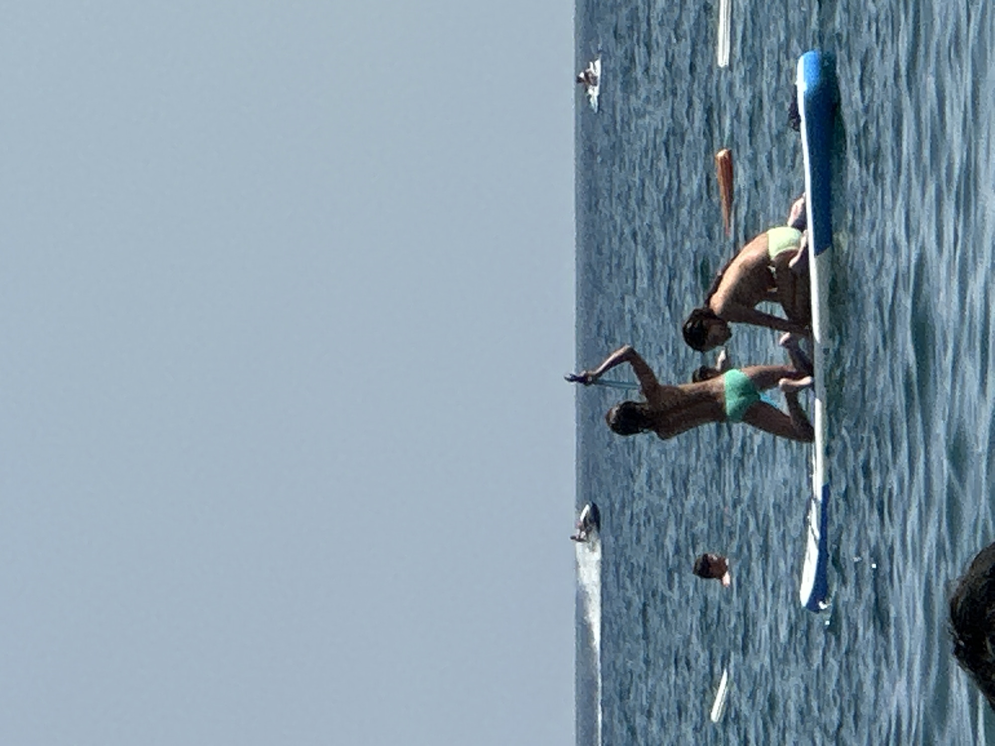 In the waters off the coast of Boreti, Montenegro, two young people are on a paddleboard. One is kneeling and appears to be holding a paddle aloft, while the other is sitting on the board, facing away from the camera. A third person is visible in the water, swimming. In the background, a jet ski is being ridden, creating a spray of water. The sky is clear and blue.
