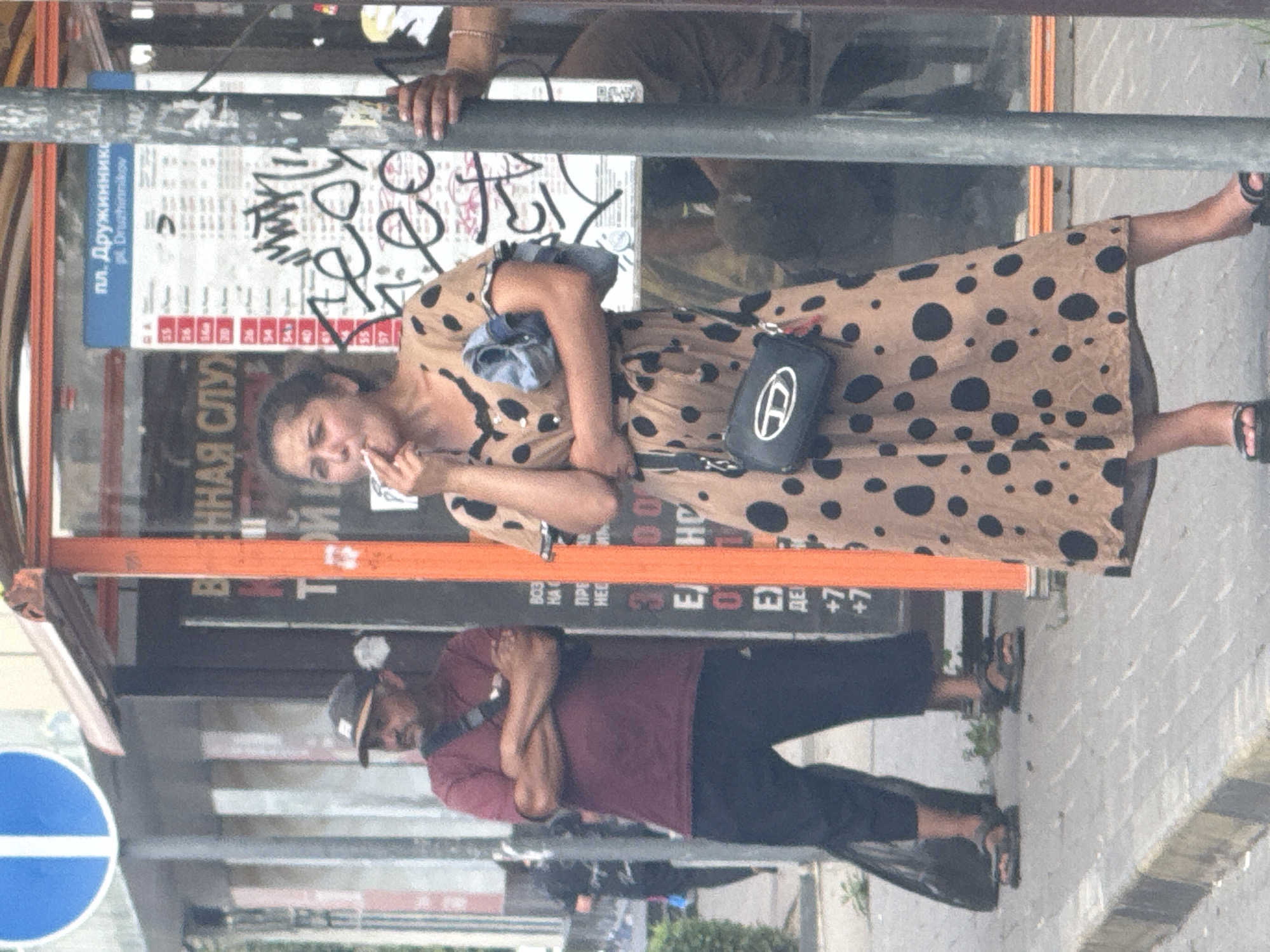 In the city of Rostov-on-Don, Russia, a man wearing a maroon t-shirt and black pants stands with his arms crossed, his gaze directed to the left. Beside him, a woman in a tan dress with black polka dots is smoking a cigarette. She holds a folded blue garment in her left arm and a black cross-body bag with a silver logo hangs from her right shoulder. They are both standing on a sidewalk in front of a bus stop shelter. Behind them, a bus schedule is displayed on a blue and white sign covered in graffiti. The environment appears to be an urban street with a pole and a traffic sign visible in the background.