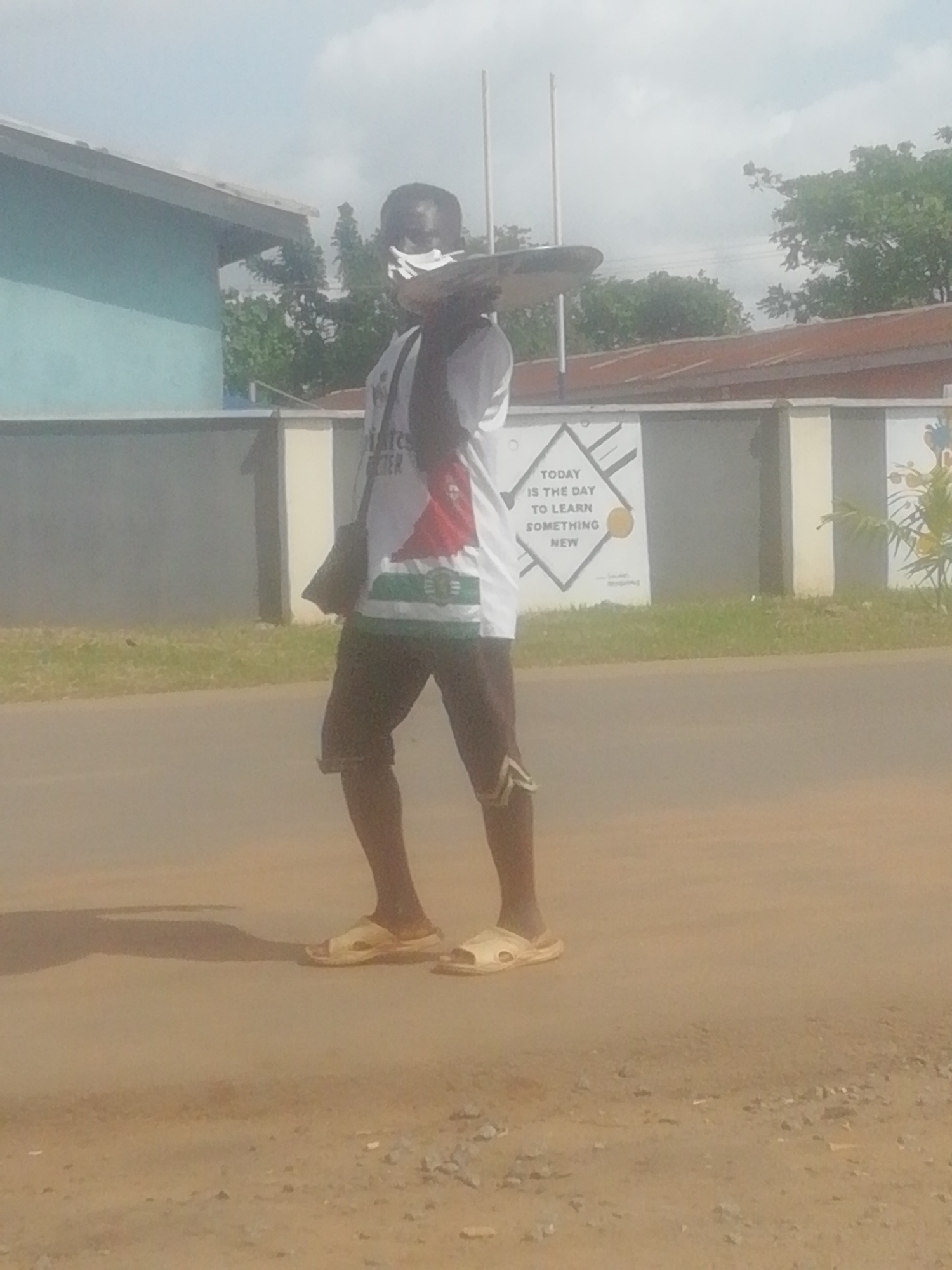 A man is walking along a road in Ikorodu, Nigeria. He is wearing a white t-shirt with the words "Pirates for Better" and a red and green design, brown shorts with a zig-zag pattern on the side, and light-colored sandals. He has a black bag slung over his shoulder and is carrying a circular object on his head. The road is dusty and appears to be unpaved in some areas. Behind the man is a wall with a mural that reads, "Today is the day to learn something new." A blue building is visible on the left side of the image, and trees are visible in the background. The sky is bright and overcast.