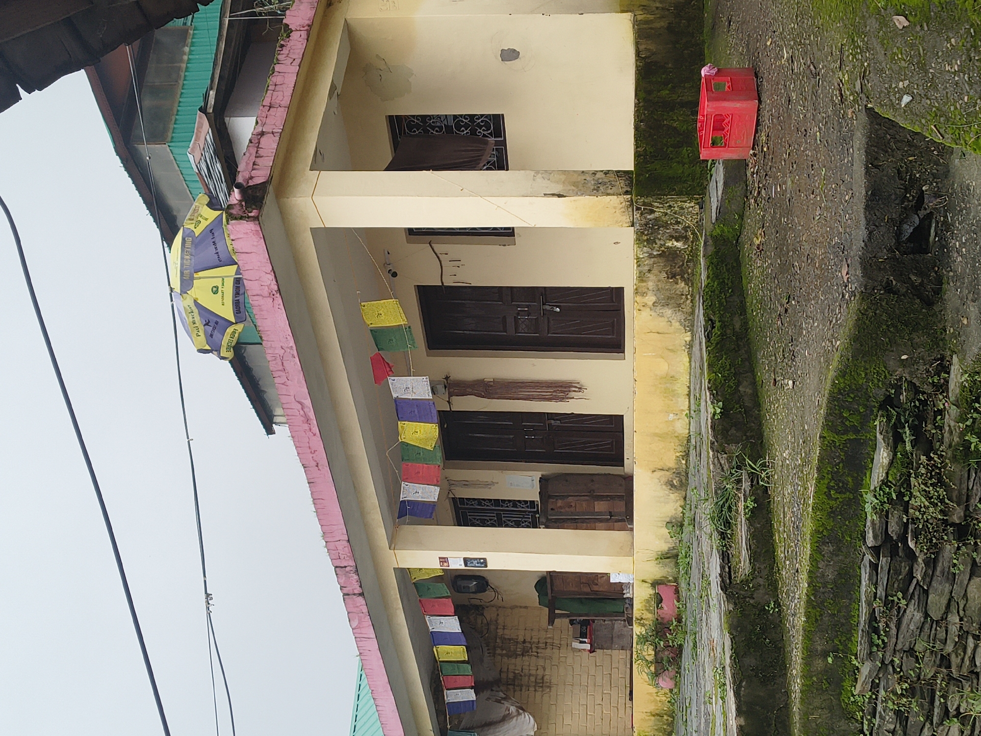 The image shows a small, light-yellow building with a pink-trimmed roof in Haini, India. Prayer flags in multiple colors are strung across the porch, which has two dark wooden doors and a window with decorative metalwork. A brown cloth hangs outside the window. Above the roof, a yellow and purple umbrella with text is visible. In the foreground, moss-covered stone steps lead up to the building, and a red plastic crate sits on the ground to the right. The overall environment appears to be overcast and damp, with vegetation growing on the steps and around the building.