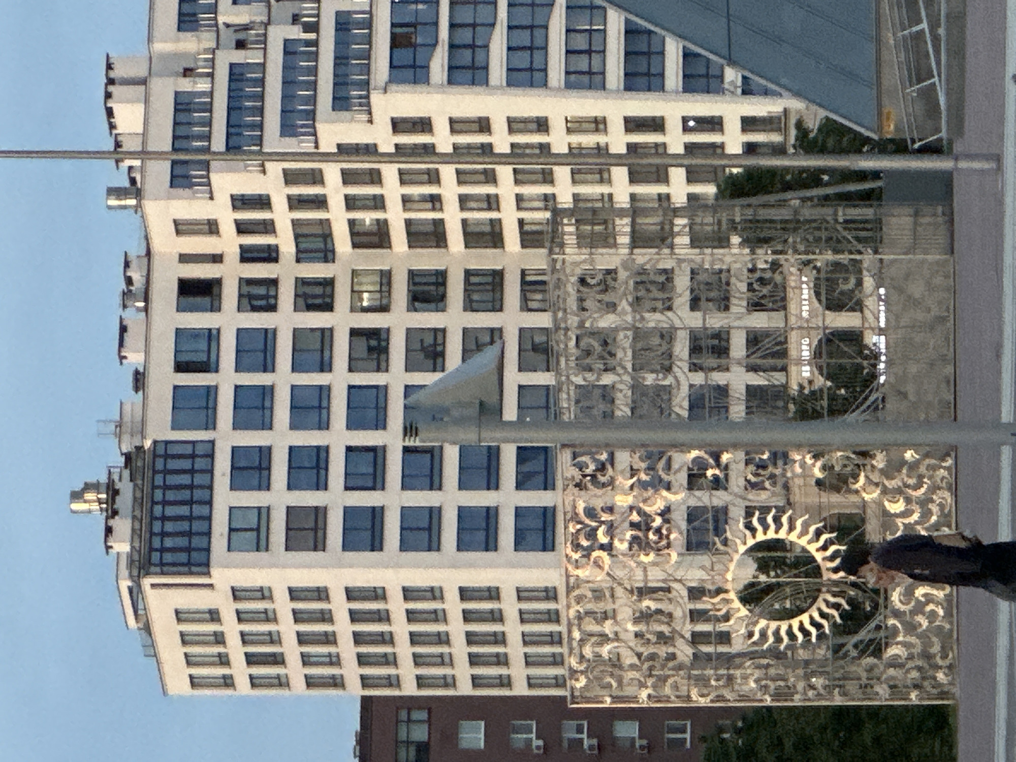 This detailed image depicts an urban scene in Minsk, Belarus, specifically near the Gallery Minsk shopping mall and Nemiga metro station, identifiable by the prominent sun-themed decorative screen and the partially visible "ЕВРО-ОПТ СУПЕРМАРКЕТ" sign behind it.
In the background, a large, multi-story building complex dominates the scene. This complex is predominantly white, featuring various window styles: one section on the left displays numerous vertically aligned, dark, rectangular windows, while another section on the right exhibits larger, square, blue-tinted windows that reflect the clear blue sky. Some sections of the building also show multiple balconies with metallic railings. The rooftops are adorned with several white, chimney-like structures and at least one prominent, cylindrical metallic vent. To the far left, a multi-story brown building with smaller, rectangular windows and visible external air conditioning units is partially obscured.
In the mid-ground, a large, ornate metal screen stands vertically, featuring intricate swirling patterns and a distinct central circular motif resembling a stylized sun with radiating rays. The metallic surface of this screen reflects sunlight, creating bright highlights. Behind and through this screen, faint reflections and partial views of other structures, including what appears to be the "ЕВРО-ОПТ СУПЕРМАРКЕТ," are visible.
In the lower foreground, a single individual is walking from left to right on a paved surface. The person is dressed in dark clothing, possibly a coat or jacket, and is carrying a light-colored bag or object in their left hand. Their face is partially obscured. To the right of the walking person, a tall, slender, light-colored pole extends vertically, partially obscuring the background. In the lower-right corner, a dark, pyramid-shaped structure with a reflective, blue-tinted surface is visible, suggesting a modern architectural element or a metro entrance.
The sky overhead is clear and light blue, indicating daytime conditions. Green foliage from trees is visible on the far left, behind the brown building and the decorative screen. The lighting suggests a bright day, casting reflections on the building windows and the metallic screen.