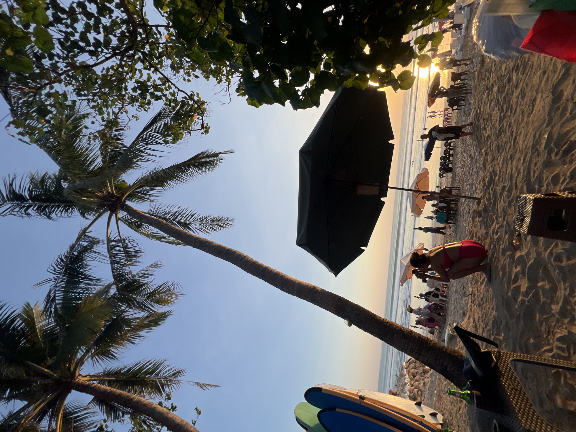 This is a vertical, eye-level, outdoor photograph taken at sunset on a beach in Kuta, Indonesia. Two prominent palm trees lean into the frame from the left and center, their fronds silhouetted against a clear, pale blue sky transitioning to a soft orange glow at the horizon. To the left, a stack of surfboards, one blue and one green, are propped up. In the foreground, a dark umbrella shades a sandy area. A woman in a pink bikini is crouched on the sand, looking towards the ocean and holding a smartphone, seemingly taking a photo or video. She is positioned to the right of a wicker-like table and chair set. Further into the background, several people are seated in chairs under umbrellas, some appearing to be at a beachside establishment. Others are walking along the beach or standing near the water, where gentle waves are visible. The sand is golden and textured, with footprints and shadows cast by the setting sun. The overall atmosphere is relaxed and tropical, characteristic of a beach scene at dusk.