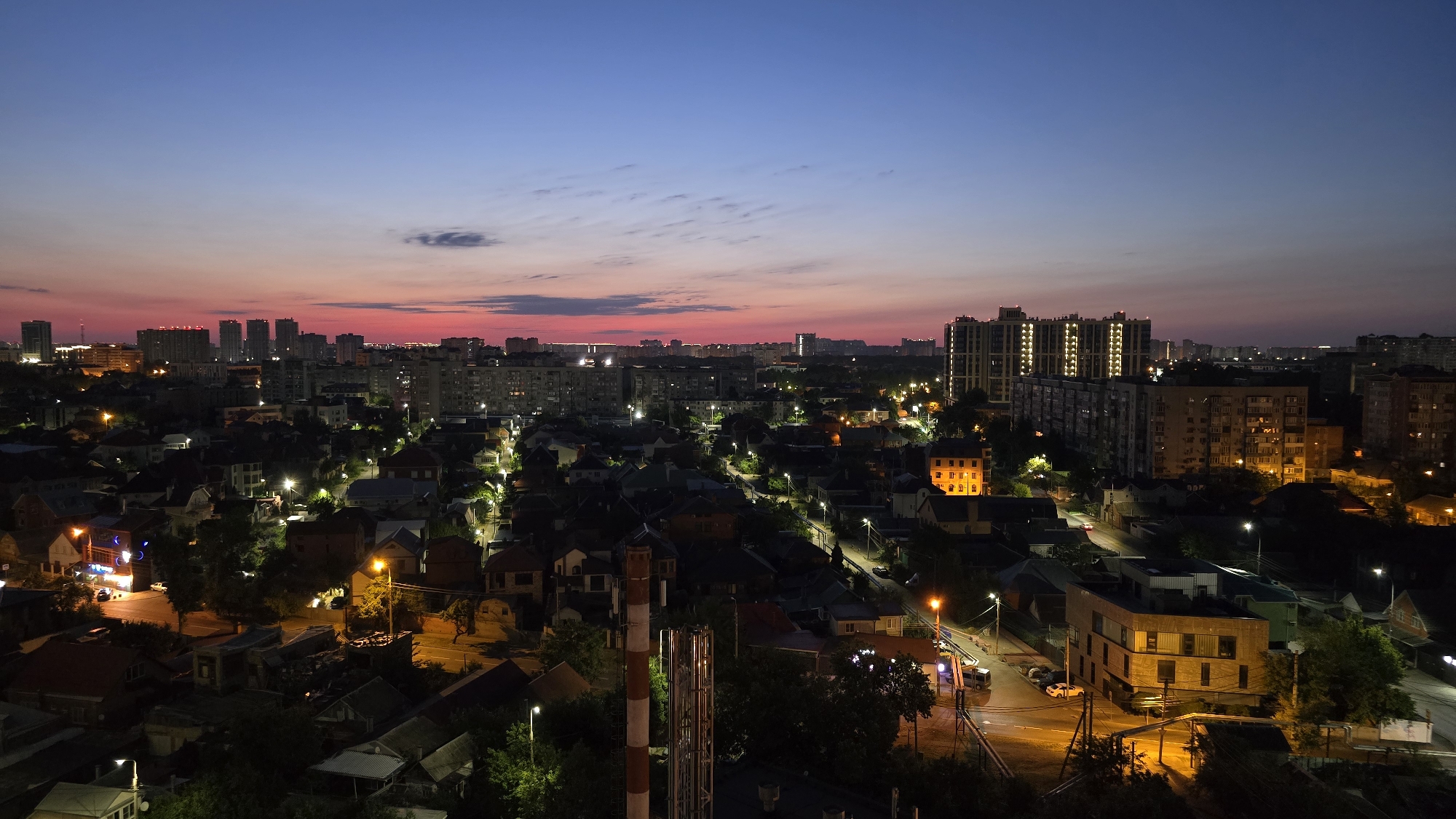 This aerial, wide-angle photograph captures a city skyline at twilight in Krasnodar, Russia. The sky transitions from a deep twilight blue at the top to a band of pink and orange hues near the horizon, indicative of sunset or sunrise. Below the sky, a dense urban landscape unfolds, characterized by a mix of mid-rise apartment buildings and lower-rise residential houses. Many buildings have illuminated windows, suggesting that it is evening. Streetlights cast pools of yellow light on the roads and illuminate the facades of some buildings. In the foreground, there's a section of what appears to be industrial piping, painted with red and white stripes, and some darker, less defined residential structures. The overall scene depicts a quiet evening in a residential and commercial district of the city.