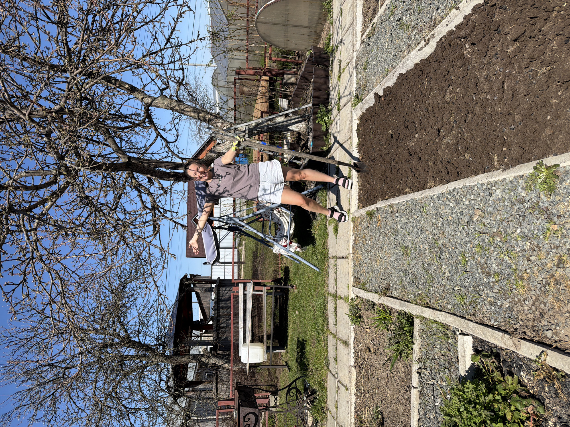 A person is standing in a garden, holding a shovel. The person has short brown hair and is wearing a gray t-shirt, white shorts, and black and white sandals. The garden has a plot of freshly tilled soil and a gravel path. In the background, there is a tree with bare branches, a metal swing set, a white plastic container, a metal grill, and a greenhouse. The sky is blue and cloudless. The location is Chaikovsky, Russia.