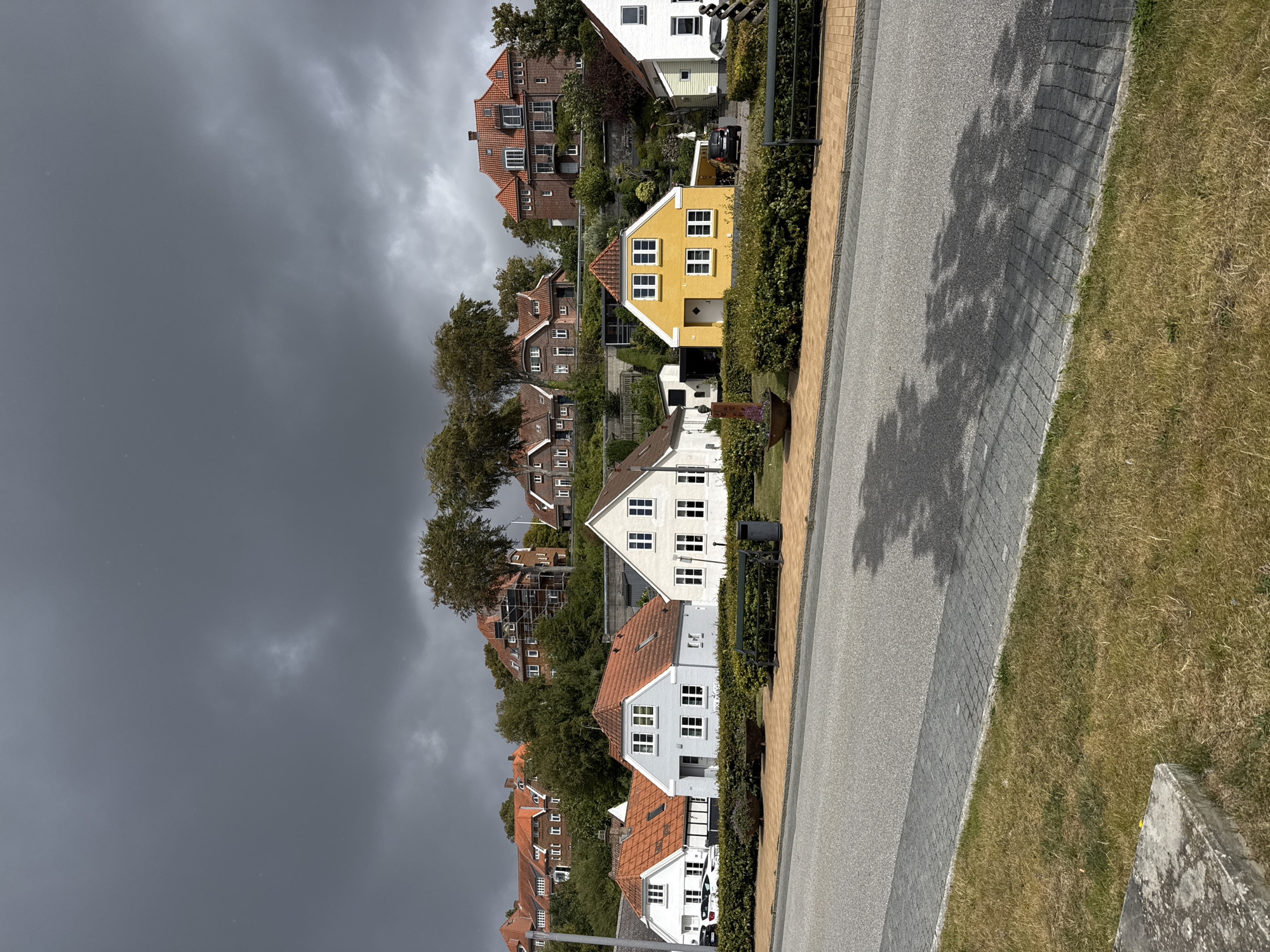 The image shows a street in Sønderborg, Denmark, with a row of houses on a hill overlooking the street. The houses are of various architectural styles, but many feature red tile roofs. Some of the houses are painted white or yellow, with white window frames. The houses are situated on a slope covered with green shrubs and trees. The sky is overcast with dark, heavy clouds, suggesting the possibility of rain. In the foreground, a paved street runs horizontally across the image, bordered by a sidewalk and a grassy area. Shadows of tree branches are cast on the street and sidewalk. There are no people visible in the image. The scene appears to be residential and tranquil, despite the ominous sky.