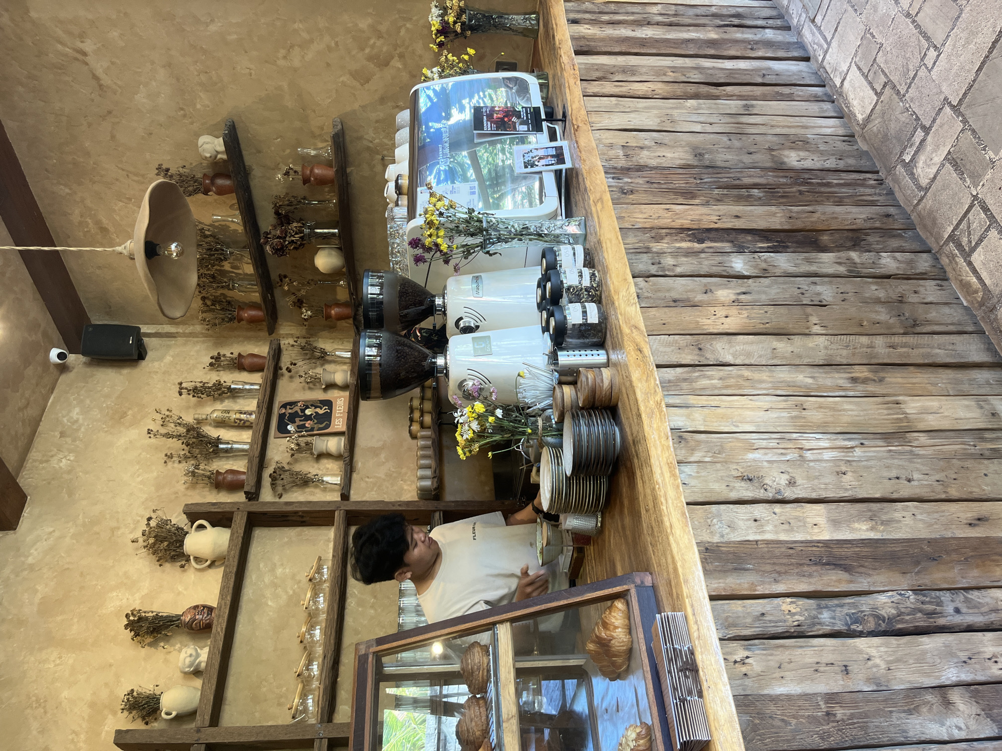 A person stands behind a coffee bar in a cafe, located in Kuta Selatan, Indonesia. The barista, dressed in a light-colored t-shirt, faces forward and interacts with items on the counter. The counter features two white coffee grinders and an espresso machine. Yellow and purple flowers in vases are placed between the machines. Behind the counter, a rustic wooden wall displays various decorative items in vases and jars. A single lamp hangs from the ceiling. A wooden floor is visible to the side. A display case with croissants sits at the edge of the counter near the barista.