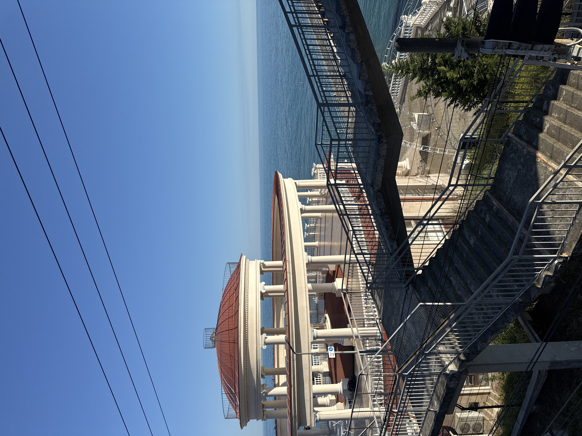 The image depicts a coastal scene with a grand rotunda structure overlooking the sea. The rotunda, with its white colonnades and reddish-brown domed roof, is a prominent architectural feature. In the foreground, a concrete staircase with metal railings descends, leading towards the rotunda and the sea. The sky is clear and blue, and the sea is calm and vast.
The location is identified as the Rotunda at the port of Sochi, Russia. This landmark is situated on the coast, offering panoramic views of the Black Sea. The architecture suggests a historical or resort-style setting. The image appears to be taken during daylight, with bright sunlight casting shadows. There are no people visible in the scene, emphasizing the architectural and natural elements. The presence of utility wires in the upper portion of the image indicates the urban context of the location, despite its scenic beauty.