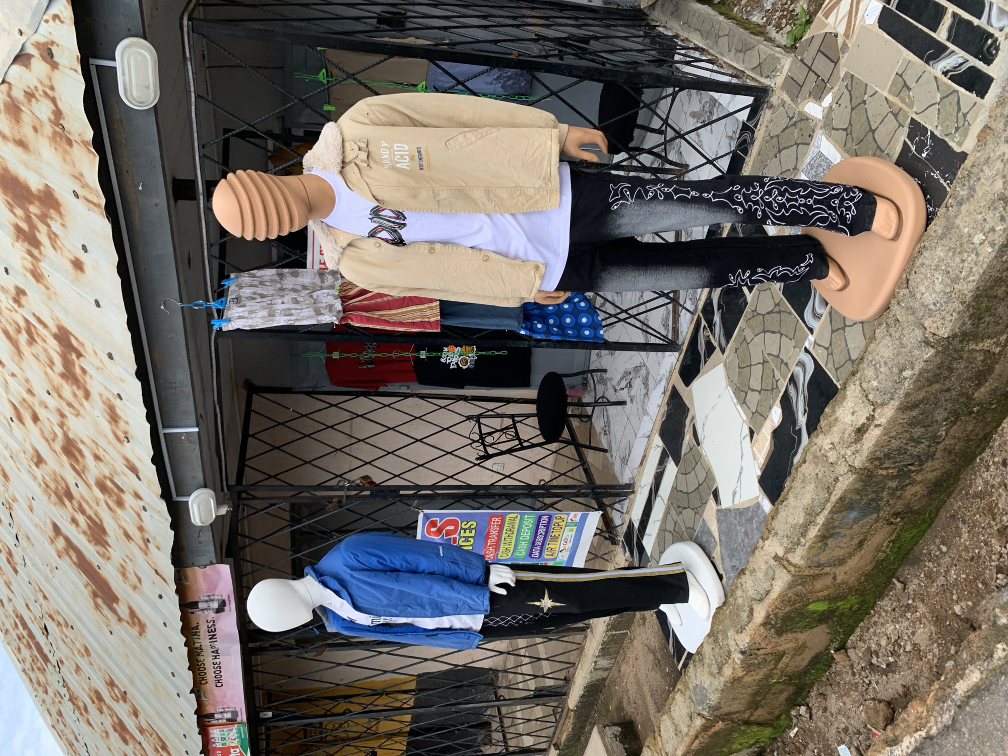 The image shows two mannequins dressed in casual clothing displayed outside a shop. The mannequins are placed on a mosaic tiled surface, with a rusty corrugated iron roof overhead. The shop appears to be a retail establishment, possibly selling clothing or related items, as indicated by the garments on display and a sign with various service offerings.
The mannequin on the left is wearing a beige jacket with a furry collar over a white t-shirt and dark, patterned pants. The mannequin on the right is dressed in a blue jacket over a white shirt and black pants with a gold stripe and star emblem. Both mannequins are wearing white footwear, possibly flip-flops or sandals.
Visible text in the image includes "CHOOSE MATINA. CHOOSE HAPPINESS." on a pink sign, and "HANDY ACID NOT INSTANTS" on the beige jacket. Another sign lists services like "CASH TRANSFER," "CASH WITHDRAWAL," "CASH DEPOSIT," "DATA SUBSCRIPTION," and "AIR TIME TOPUP." The scene suggests a daytime setting, with no specific weather conditions immediately apparent. The overall impression is of a small business display in an urban or semi-urban environment, possibly in Jos, Nigeria, given the provided location context.