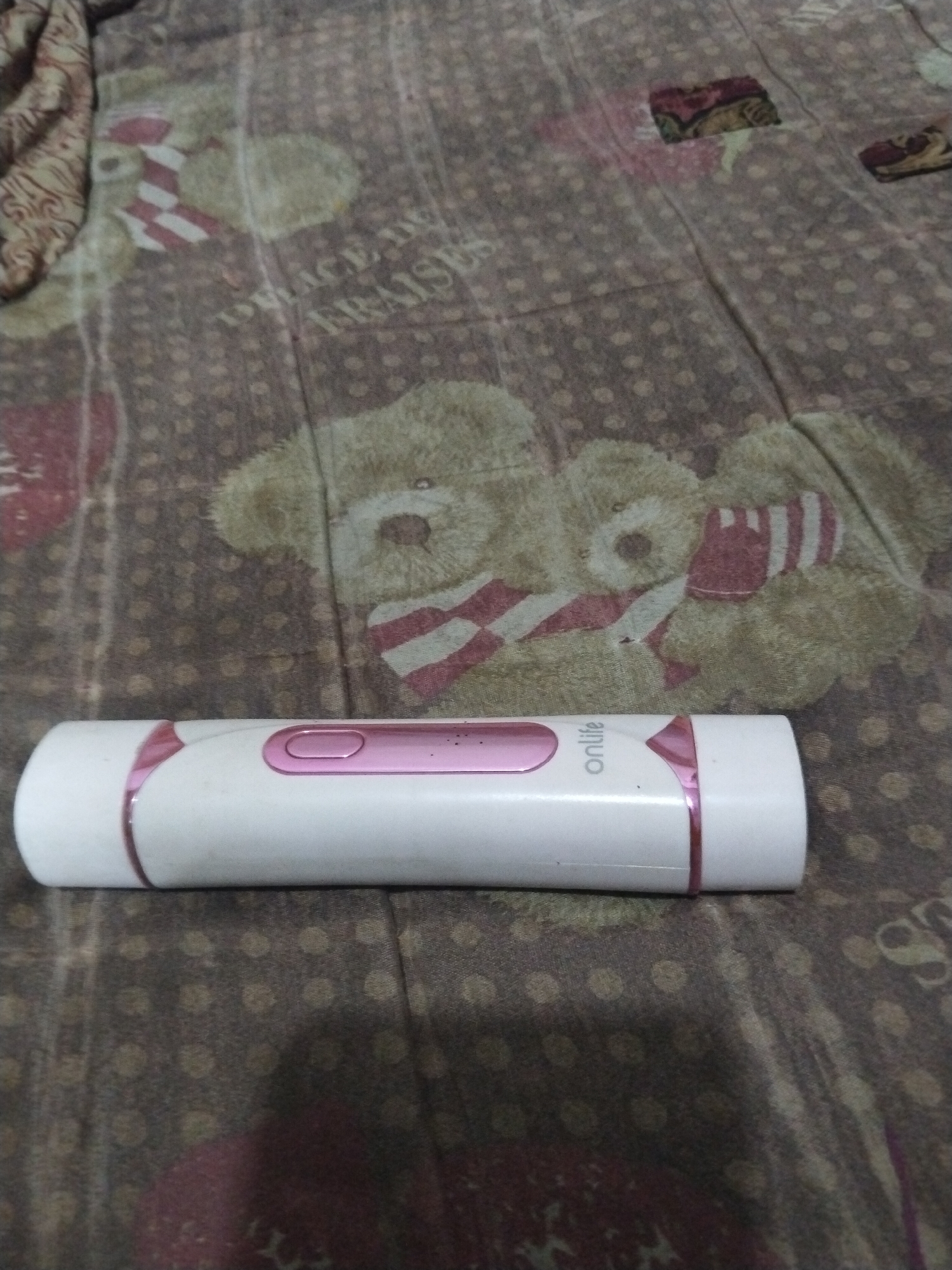 A white cylindrical electronic device with pink accents lies horizontally on a brown patterned fabric. The fabric features a repeating pattern of teddy bears wearing striped scarves and small polka dots. The device has a pink oval-shaped button and the word "onlife" is printed in white text on its surface. The overall scene appears to be indoors, possibly on a bed or sofa, given the fabric. The lighting is dim and casts a slight shadow on the fabric. No people are visible in the image.