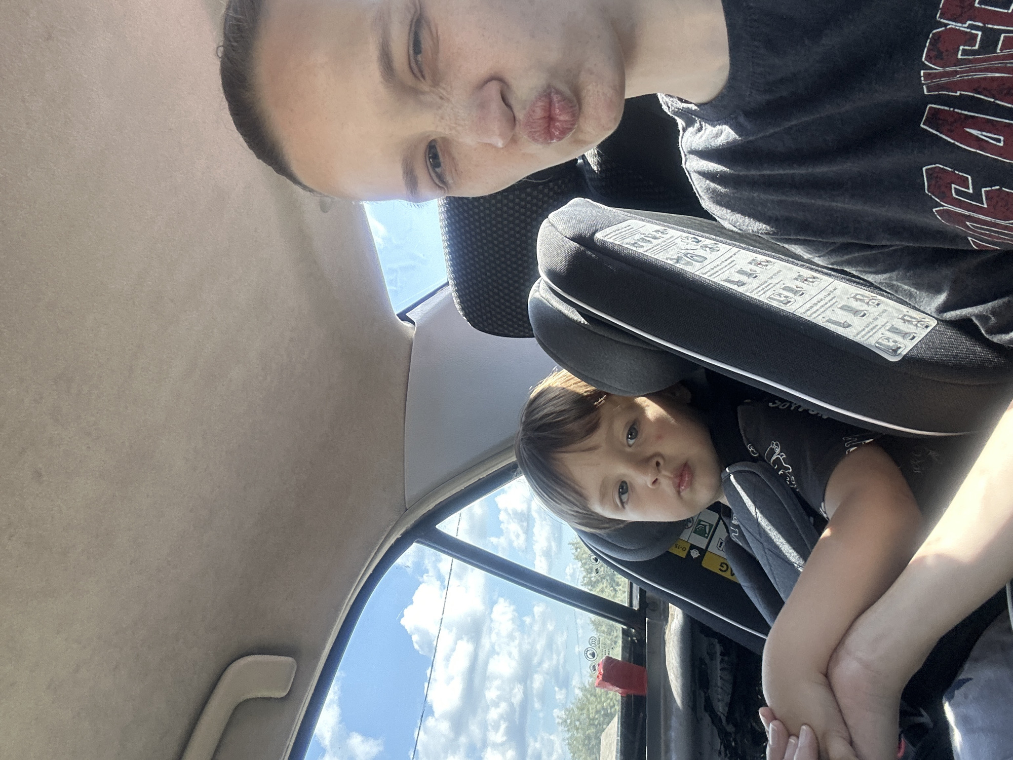 In the city of Peresyp, Russia, a selfie is taken from inside a car. A woman with fair skin and freckles, wearing a dark t-shirt with the words "LOS ANGELES" emblazoned on it, puckers her lips for the camera. To her left, a young boy with light brown hair sits in a car seat. He is wearing a dark t-shirt with a graphic design and has his arms crossed. The car's interior is visible, with a grey headliner and a grab handle. Through the car window, a bright blue sky with fluffy white clouds and distant trees can be seen. A red object is visible on the car's back seat.