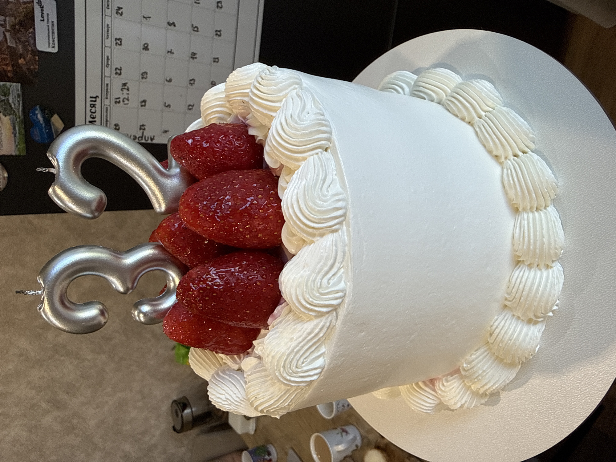 Here is a forensic description of the image:
The image shows a birthday cake with white frosting, topped with fresh strawberries. Two silver number candles, a "3" and a "2", are inserted into the strawberries. The cake sits on a white plate. In the background, a calendar with some Cyrillic text and numerals is partially visible, possibly affixed to a refrigerator. The calendar includes the names "Константин" and "Levotiip", suggesting the possibility of a personal connection. Below the cake are several white cups. Based on the calendar details, it is possible that this photograph was taken in or around Krasnoyarsk, Russia.