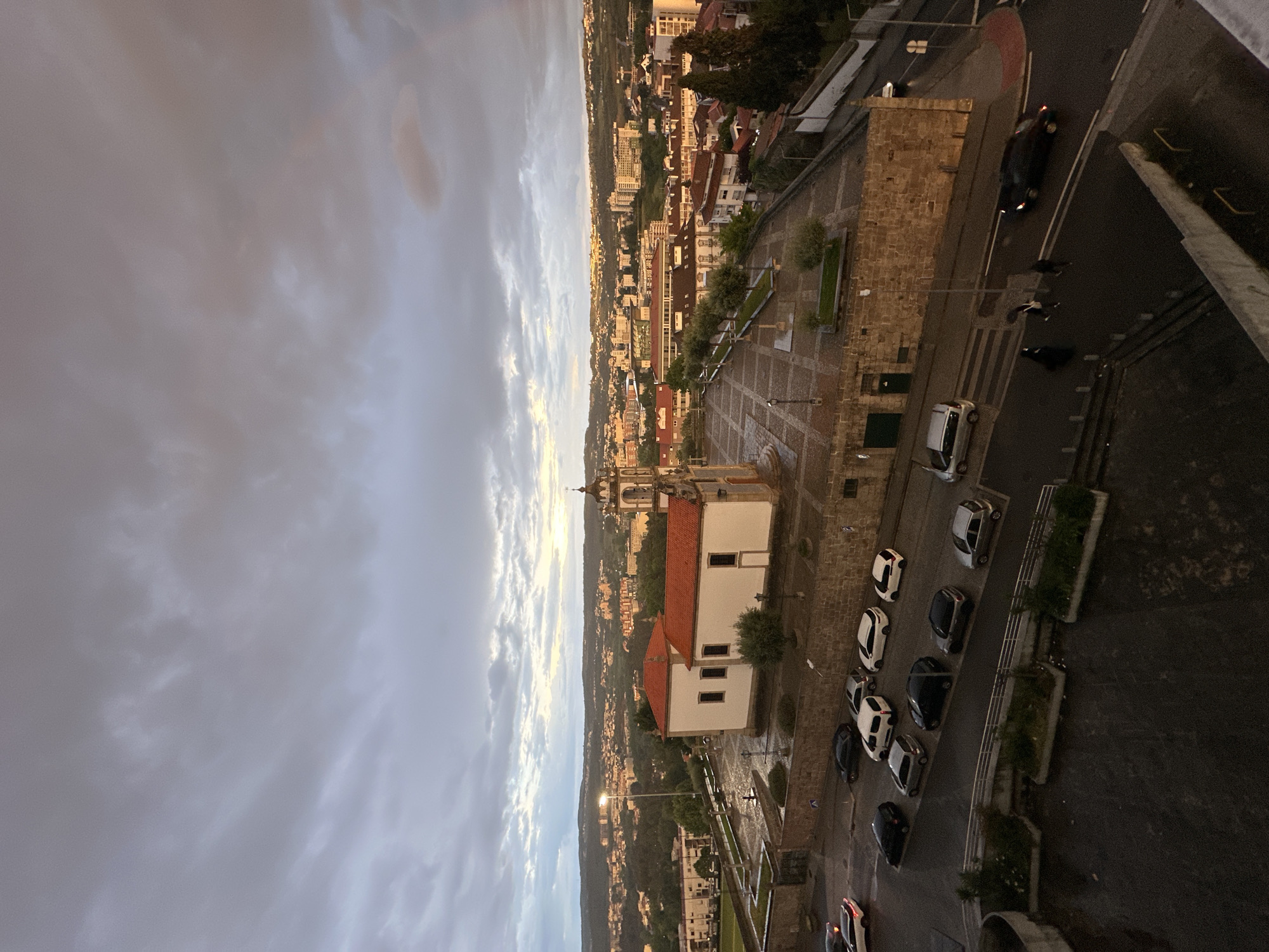 An aerial view captures a townscape at dusk or late afternoon, looking down from a high vantage point. The sky, occupying the top portion of the image, is overcast with a mix of grey clouds, some lighter patches indicating the low sun, and a warmer hue in the distance where the sun's light catches the horizon.
In the mid-ground, a prominent white building with a distinctive red tiled roof stands out, appearing to be a church or similar institutional building, complete with a bell tower and clock on its left side. Adjacent to it is a spacious, patterned plaza or square made of paving stones. Beyond this central area, the town stretches into the distance, revealing numerous buildings with characteristic red and orange tiled roofs, interspersed with trees and green spaces. The architecture suggests a European, likely Portuguese, setting, aligning with the provided location context of Vila Real.
The foreground and right side of the image are dominated by streets and parked vehicles. Numerous cars, primarily white, black, and silver, are neatly parked along a curb. One black car on a higher street section has its brake lights illuminated, suggesting recent movement or a stop. Further down, a few dark figures, possibly pedestrians, are visible on the road. The streets appear to be asphalt, with some stone wall structures lining the edges. No specific activity or event beyond everyday town life is evident. No discernible text is visible.
