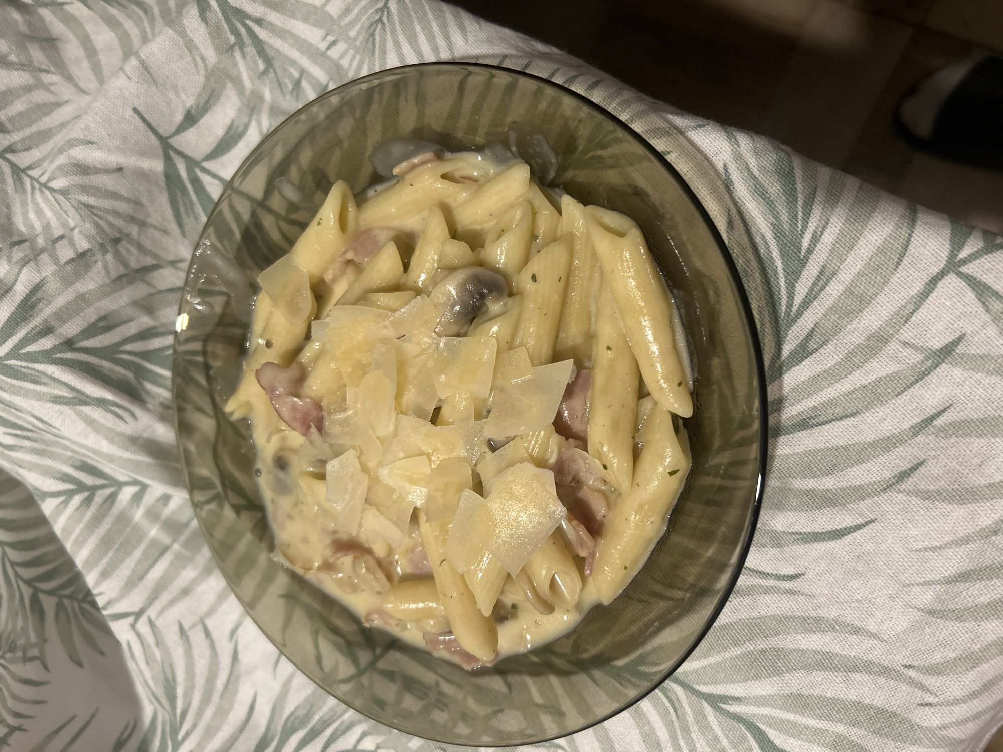 A bowl of pasta carbonara is centered in the frame, set against a patterned fabric. The pasta is topped with shavings of cheese, and pieces of ham and mushrooms are visible within the creamy sauce. The bowl is made of clear glass with a dark rim. The patterned fabric has a white background with green stylized leaf patterns. The location is Unknown, Unknown.
