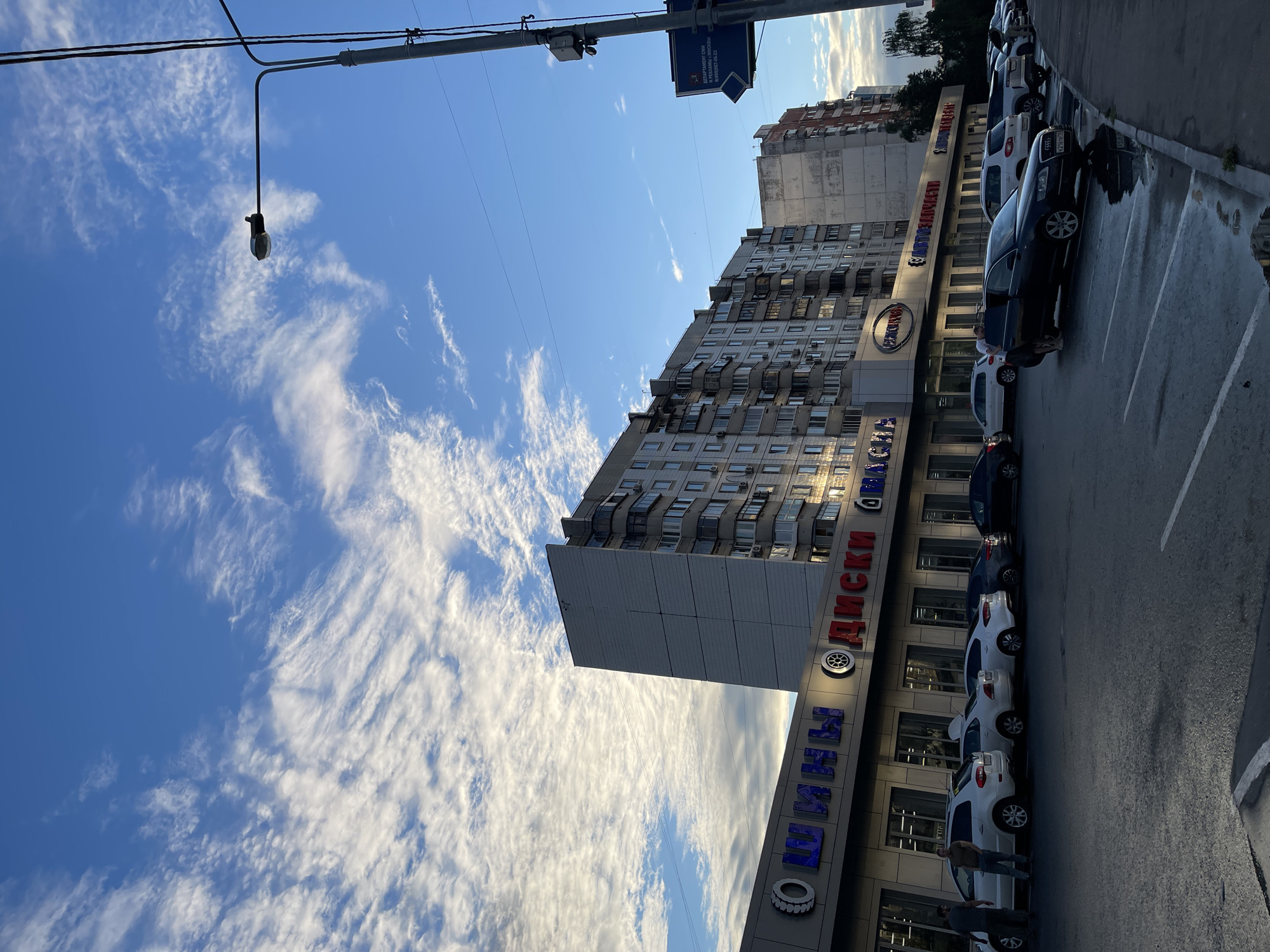 This is a medium shot, vertically oriented photo taken from a low angle. The scene depicts a street in Moscow, Russia, with a modern apartment building on the left and a commercial building with a parking lot on the right. The sky is a bright blue with scattered, wispy white clouds, suggesting it is daytime, likely late afternoon or early evening due to the soft lighting and shadows.
The apartment building is tall and contemporary, featuring many windows and balconies. Adjacent to it is a commercial building with a prominent sign displaying Cyrillic text that appears to be related to car tires and wheels, as indicated by "ШИНЫ ДИСКИ" (Tires Wheels) and a tire symbol. Below this, another sign reads "ДЕПАРТАМЕНТ СМИ И РЕКЛАМЫ МОСКВЫ" (Department of Mass Media and Advertising of Moscow). Several cars are parked in a lot in front of the commercial building, including white SUVs and a dark sedan. One man is visible near the dark sedan, appearing to be interacting with it or a passenger. A streetlamp hangs from wires overhead, and more power lines stretch across the sky. The overall atmosphere is urban and everyday, with a sense of a typical street scene in a Russian city. The wet asphalt suggests recent rain.