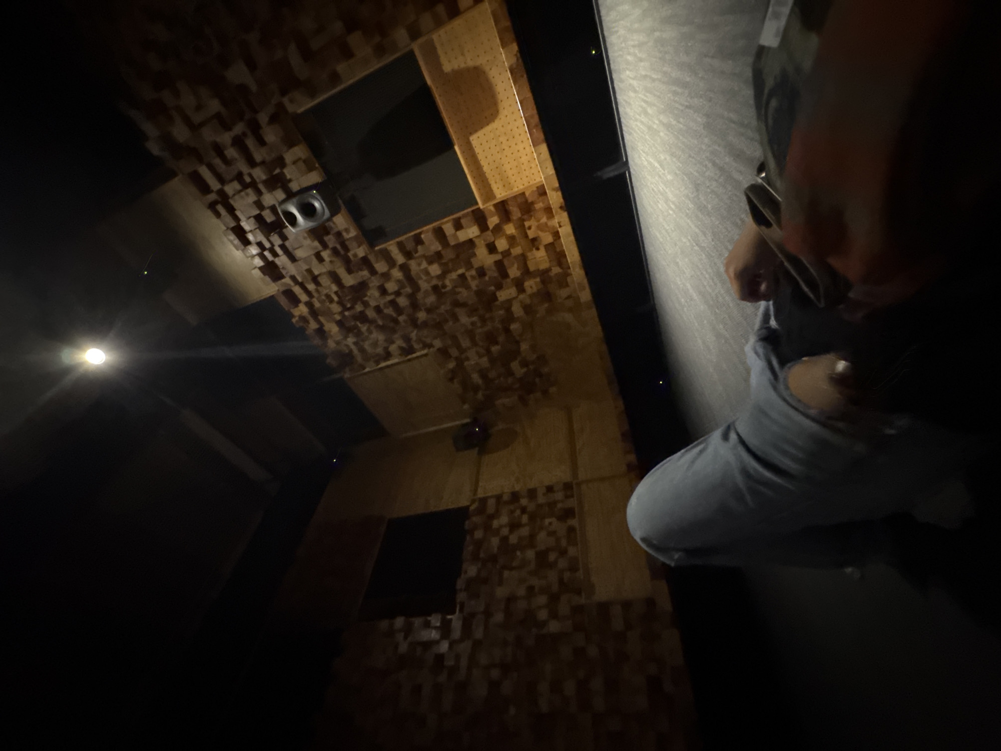 A person wearing light blue jeans and a grey sweatshirt lies on their back on the floor of a dimly lit recording studio. The studio has dark walls and ceiling, with acoustic paneling on the walls made of wood blocks. A spotlight shines down from the ceiling, casting a bright light. There is a speaker mounted on the wall. The person's hands are visible in the foreground, one holding a phone. The floor appears to be carpeted. The overall impression is one of relaxation or contemplation within a professional audio environment.