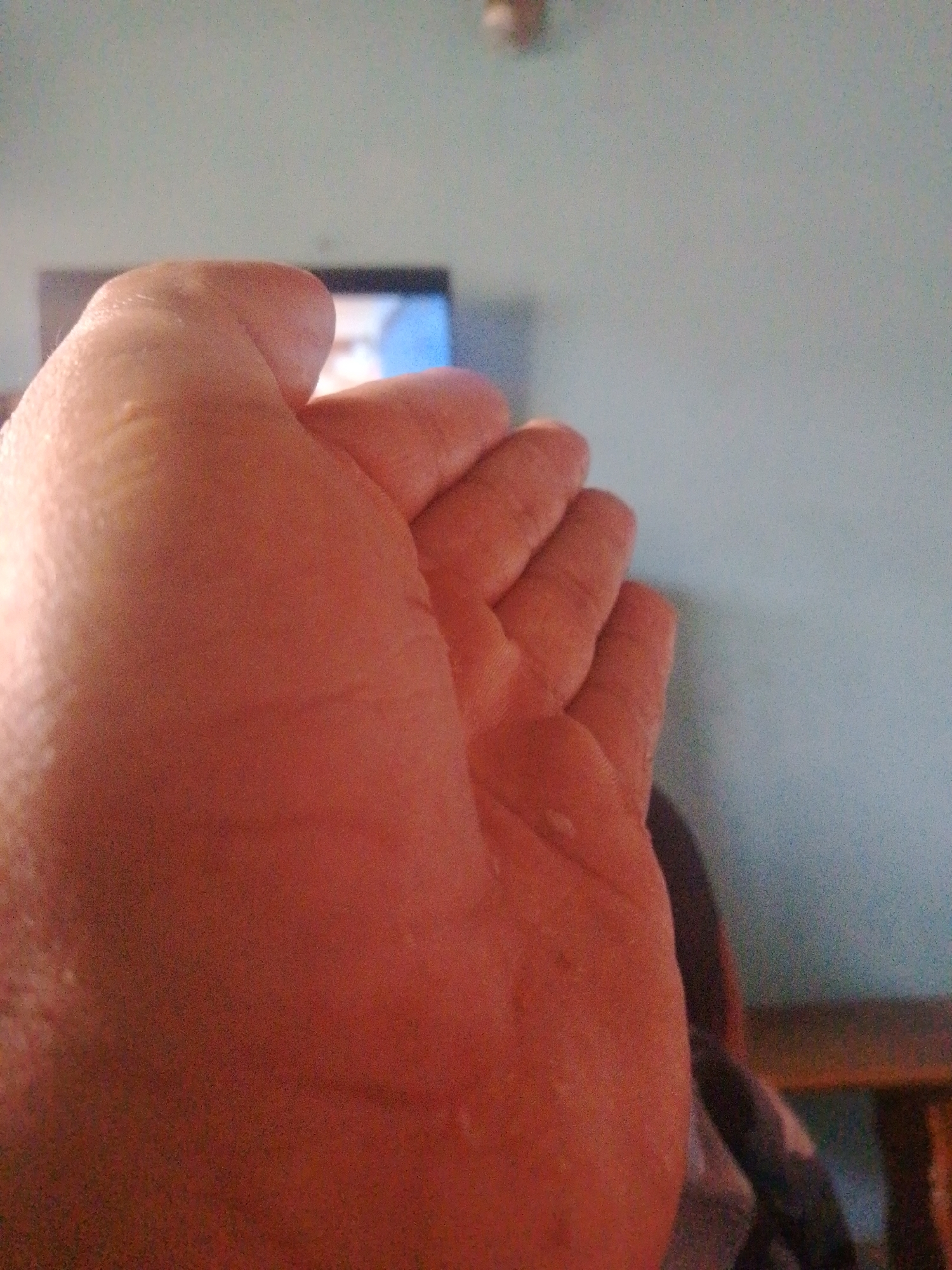A close-up photograph depicts a human hand against a blurred background. The hand is light-skinned and fills much of the frame. In the background, a television screen with unclear imagery is visible, along with a blue wall. The presence of a partially visible chair suggests an indoor setting. No specific details indicate a location in Juan Griego, Venezuela, making it impossible to confirm the location based solely on the visual information provided.