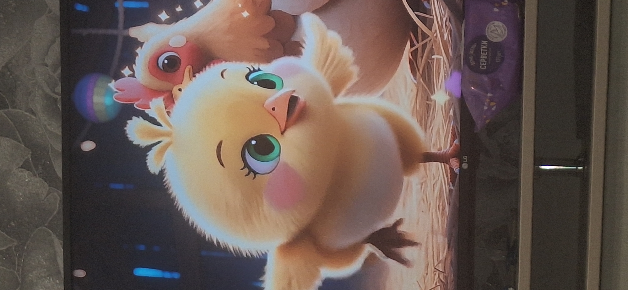 A television screen displays an animated scene with two cartoon chickens. The foreground features a fluffy yellow chick with large green eyes, pink cheeks, and its wings spread. Behind it, a larger chicken with a red comb and similar large eyes is partially visible. The background appears to be a dimly lit barn interior with some glowing elements and a colorful, striped sphere. Below the screen, a portion of a purple package, likely containing wet wipes, is visible with Russian text that translates to "WET WIPES". The logo of LG is also visible on the television frame. The image does not depict any specific location in Bila Tserkva, Ukraine.