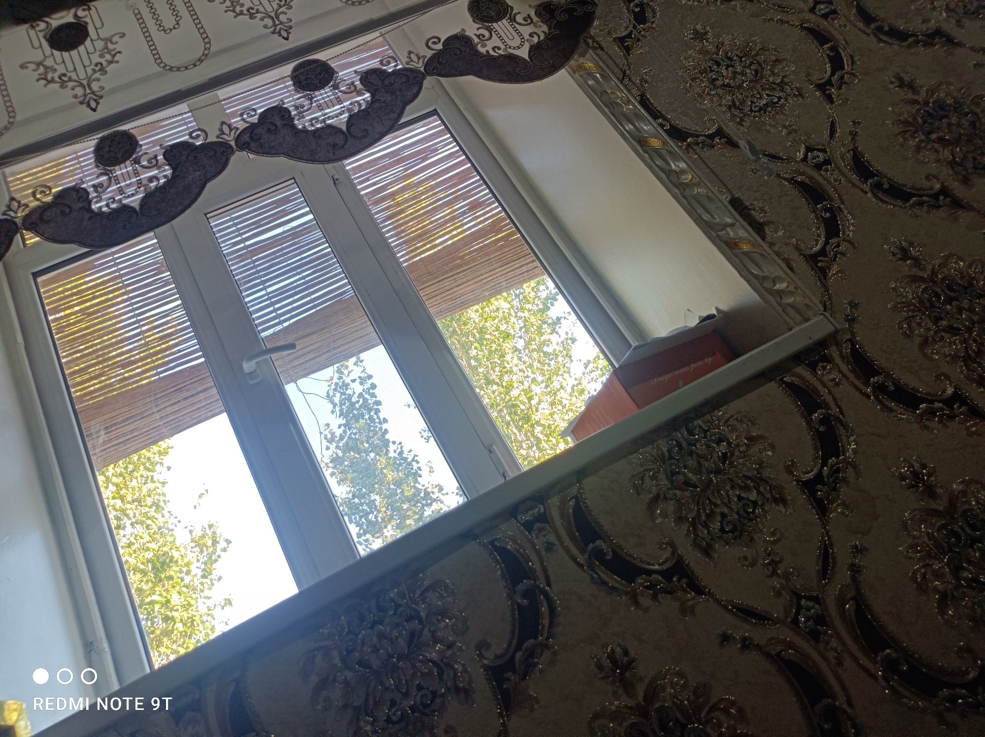 The image shows a close-up of a window with blinds and patterned wallpaper. The window is a white vinyl frame with three panes. Inside the window, there are bamboo blinds. Outside the window, green trees are visible against a bright sky. The wallpaper on the right side of the image has a damask pattern in beige and dark brown. The windowsill appears to be white. A small orange box is visible on the windowsill on the far right. The bottom left corner of the image has a watermark that reads "REDMI NOTE 9T" with circular icons. The scene is an interior view looking out of a window in a room.