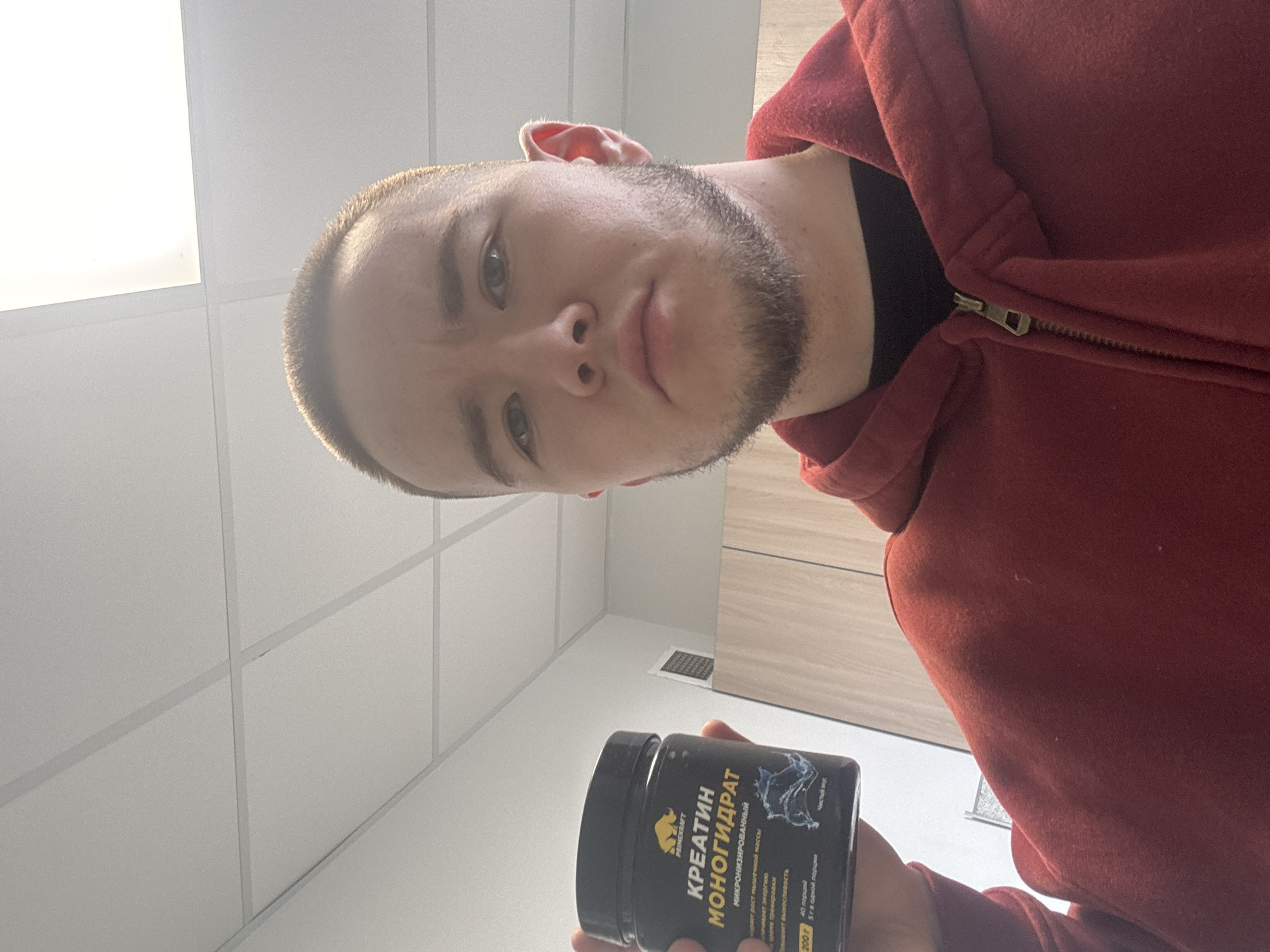 A man is holding a container of PRIME KRAFT Creatine Monohydrate supplement. He is wearing a red zip-up hoodie. The setting appears to be indoors, with a tiled ceiling and light-colored walls. A vent is visible in the wall. The product label is in Russian. The exact location is not ascertainable from the image.