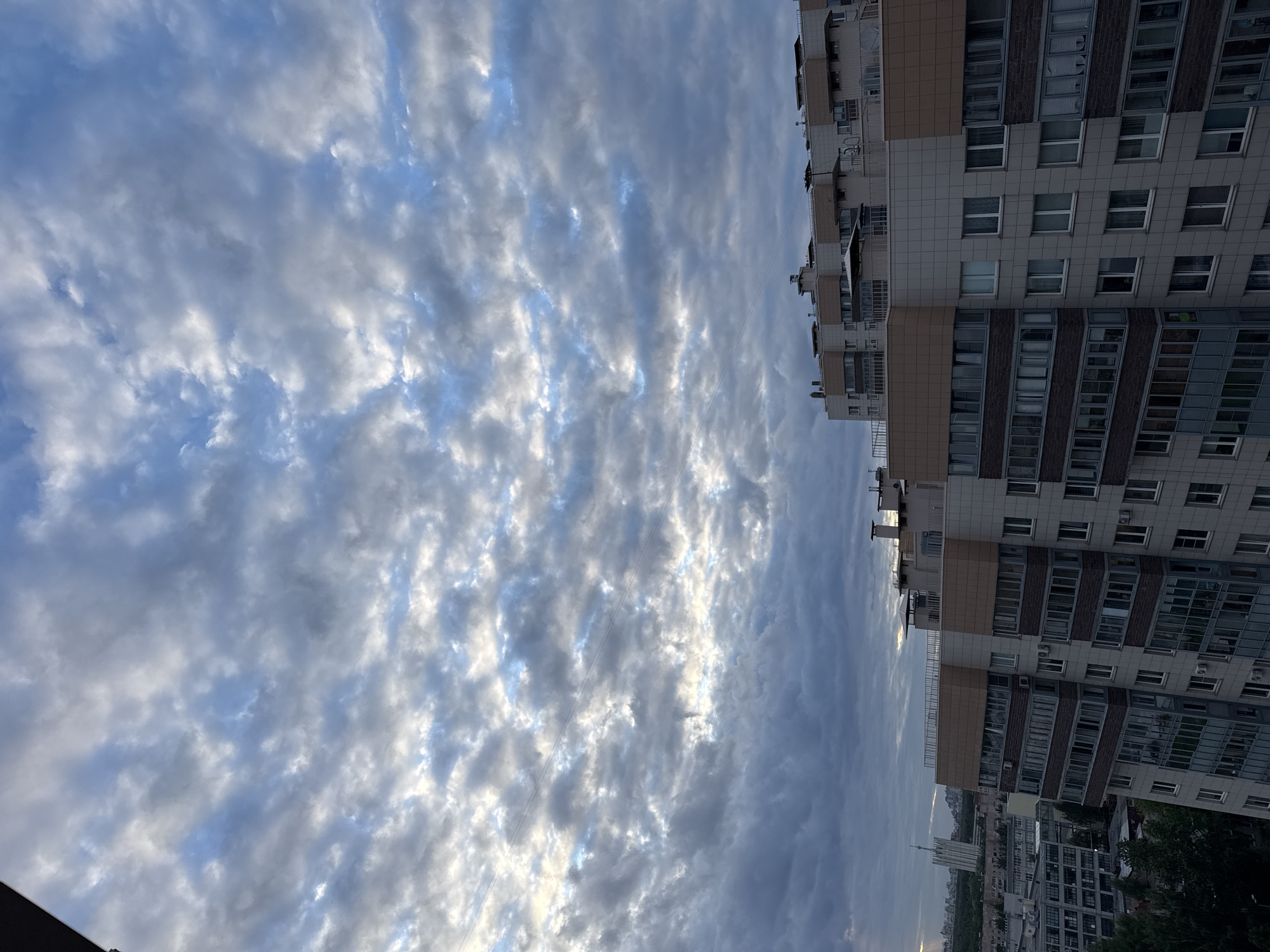 The image shows a sky filled with altocumulus clouds in a linear pattern, with white and gray tones. The sky is contrasted by a view of a multi-story residential building on the right side. The building has multiple windows and balconies. Below the building, a cityscape can be seen with other buildings and some greenery. In the distance, a tower is visible. The location is Unknown, Russia.