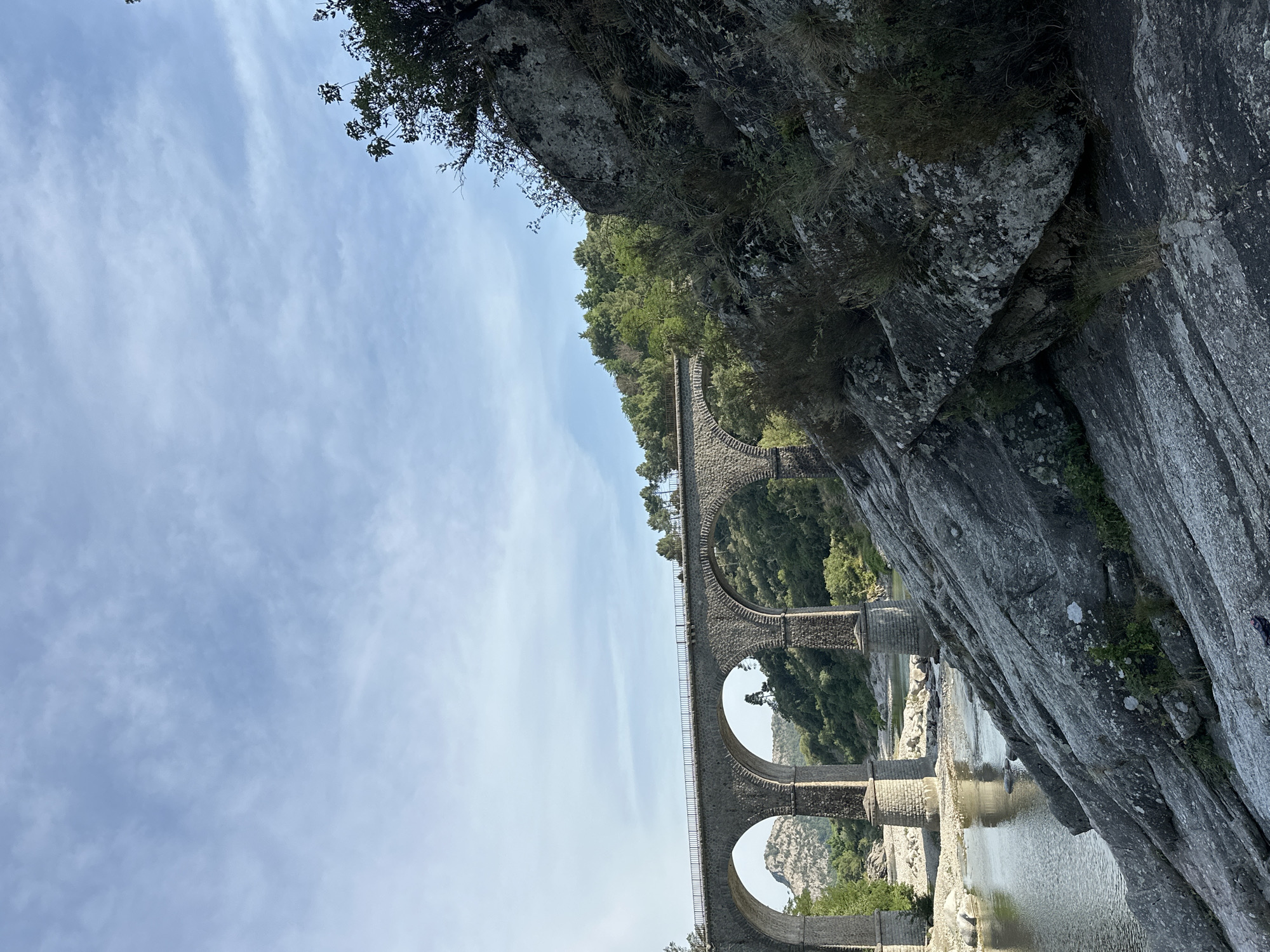 An old stone viaduct with multiple arches spans a wide, calm river. The viaduct is made of rough-hewn gray stone blocks and has a metal railing along its top. The riverbanks are rocky and lined with green trees and vegetation. In the foreground, a large, rough-textured rock formation dominates the right side of the frame, with sparse dry grass and smaller plants growing out of its crevices. The sky is a pale blue with scattered white clouds. The scene is set in a natural, rural environment, likely during daytime.