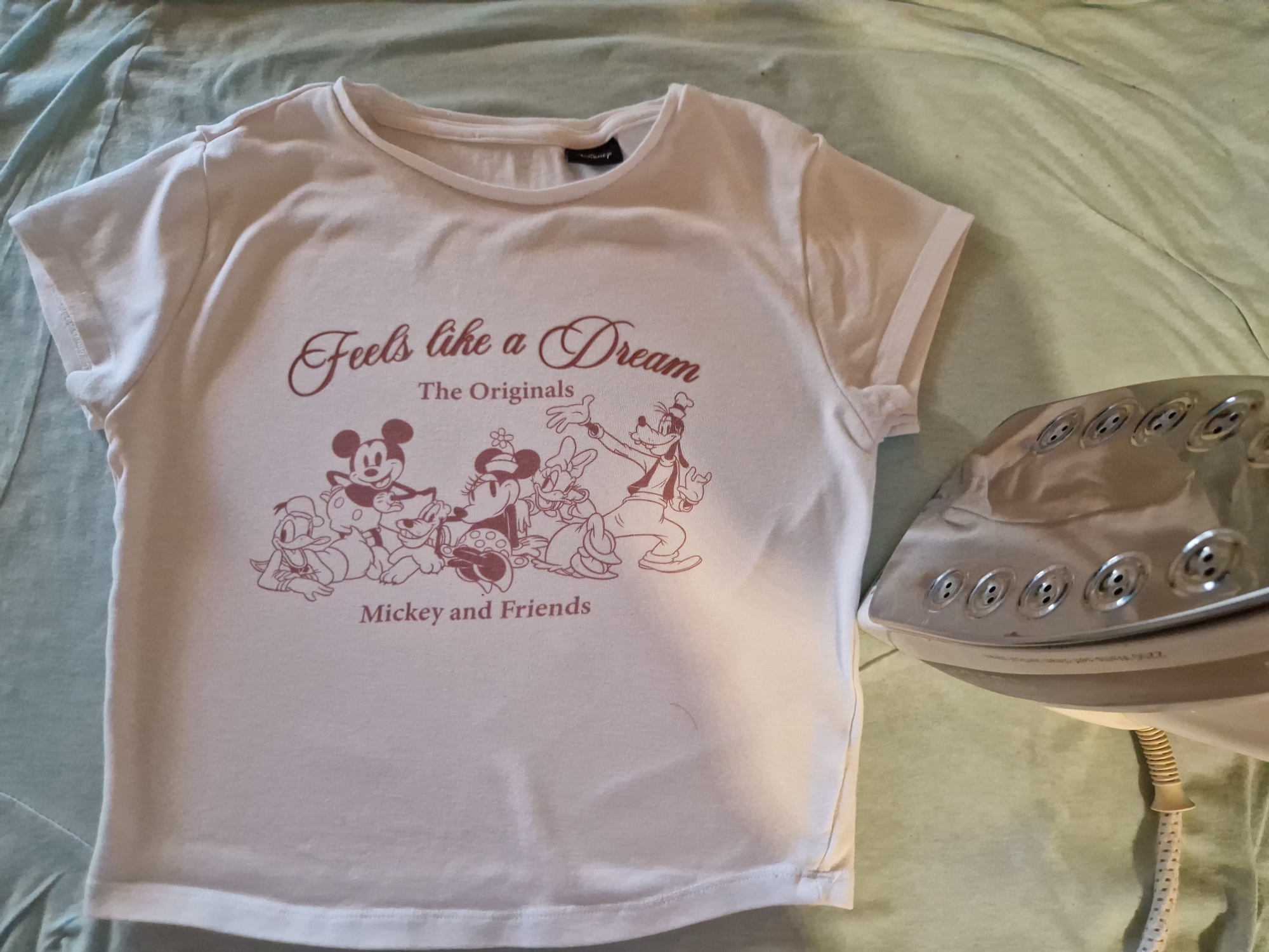An iron is resting on a light green fabric. A white t-shirt with a graphic design of Mickey Mouse and friends is draped on top of the fabric, positioned to the left of the iron. The graphic on the t-shirt features the text "Feels like a Dream" and "The Originals Mickey and Friends" in a dark pink, cursive font. The t-shirt appears to be laid out flat, possibly for ironing. The scene is indoors.