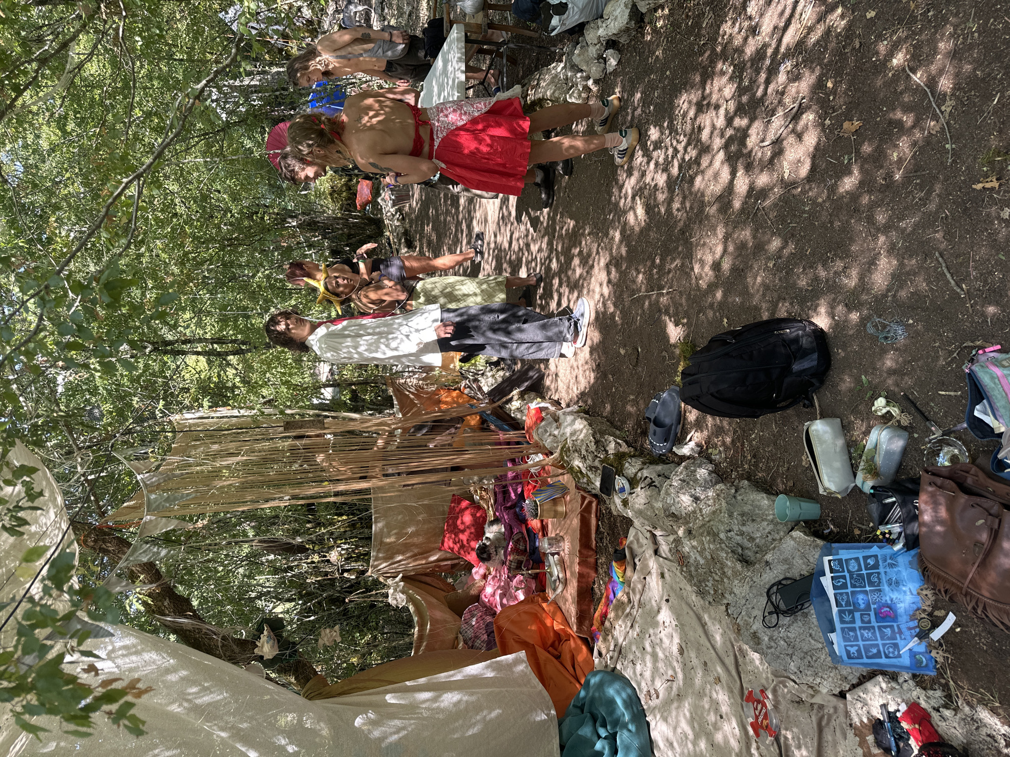 In Bukovik, Montenegro, a group of individuals gathers in a wooded area. A makeshift shelter, constructed with draped fabrics and hanging decorative elements, is visible on the left. Inside the shelter, the ground is covered with various colored blankets, pillows, and small objects. Outside the shelter, people are interacting and conversing in a clearing. The ground is covered with dirt and fallen leaves, and sunlight filters through the surrounding trees. Several bags and personal items are scattered on the ground near the shelter.