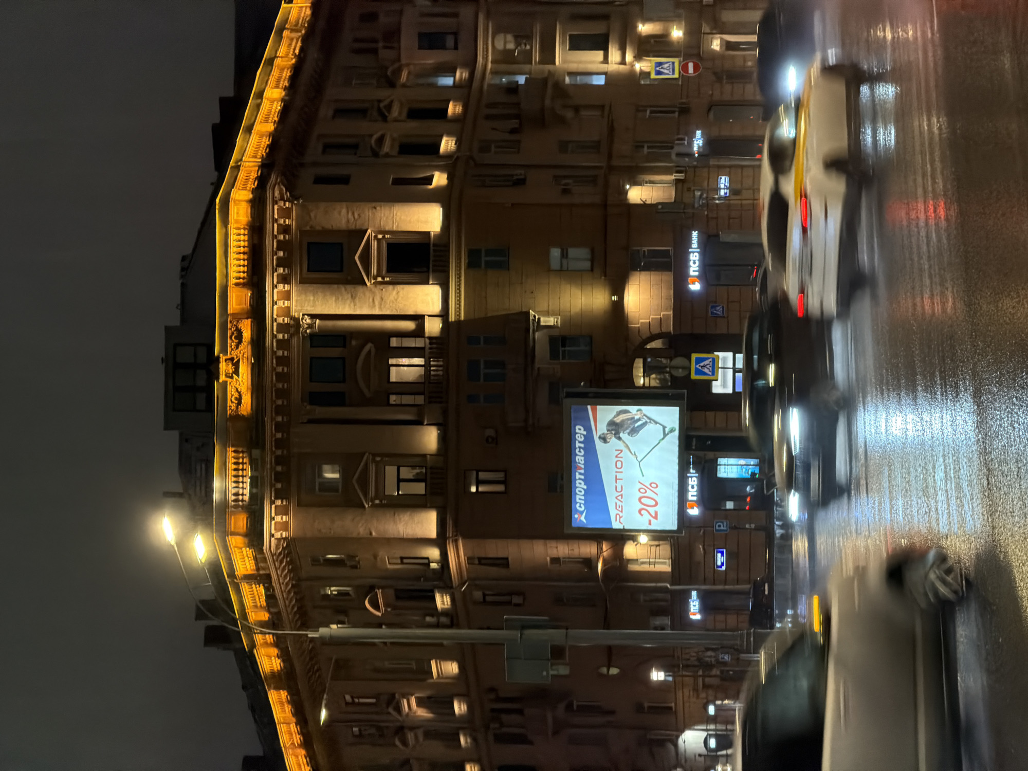 The image captures a nighttime scene in Moscow, Russia, likely along a street lined with historic buildings. The architecture of the building in the foreground features ornate details and is illuminated with warm-toned lighting, accentuating its facade. A large advertising billboard with a discount promotion is visible on the building. In the foreground, the wet asphalt street reflects the city lights and the headlights of passing cars, indicating a recent rain or drizzle. The blurring of the vehicles suggests movement, and a mix of stationary and moving cars are present. The overall atmosphere is urban and atmospheric.