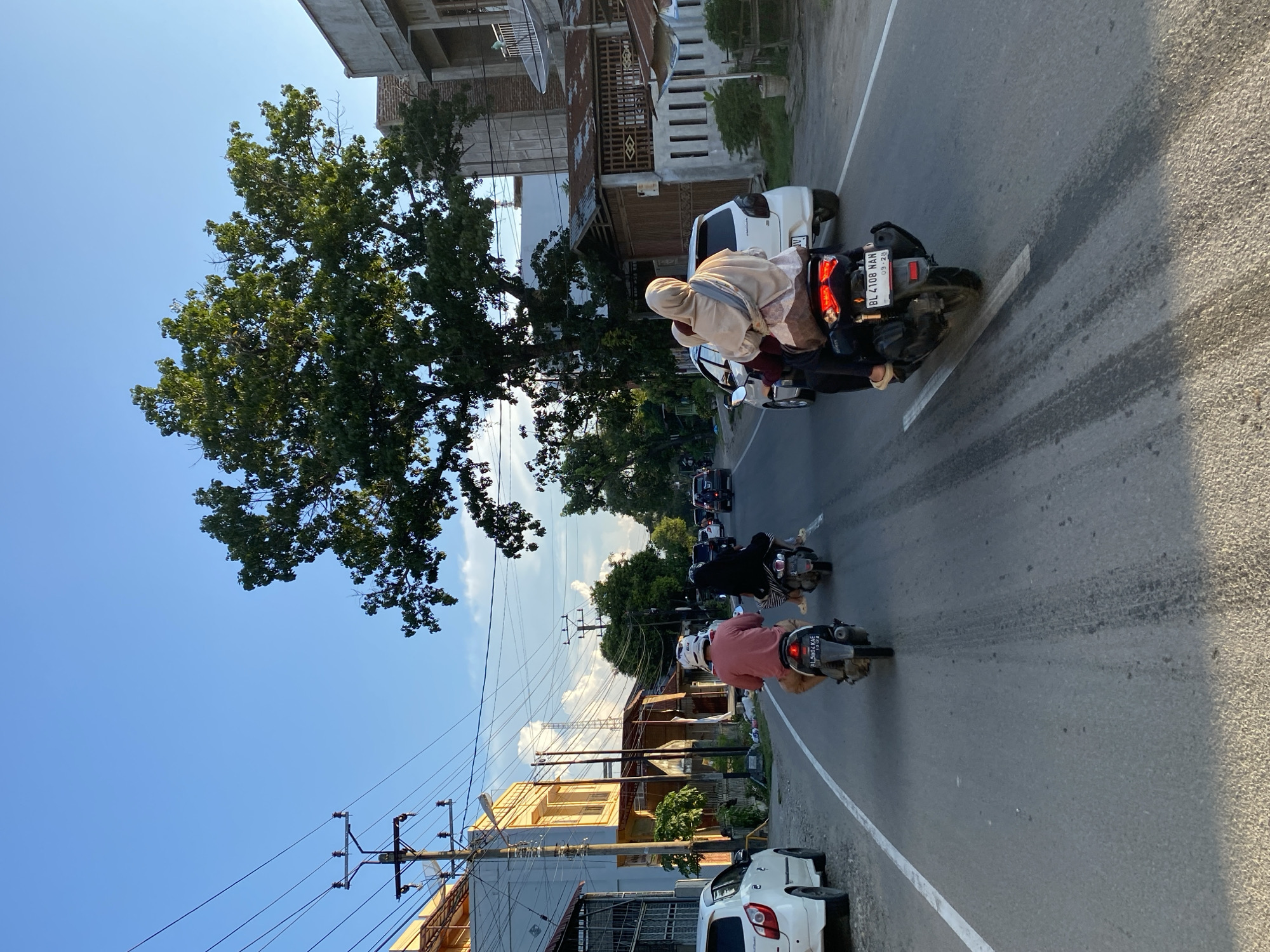 This is a ground-level, eye-level shot of a street scene in Lhokseumawe, Indonesia. The image captures a moderately busy road with several vehicles and pedestrians. In the foreground, two motorcycles are prominently featured, traveling in the same direction. The first motorcycle, a dark scooter, carries two individuals, a driver and a passenger, both wearing modest Islamic attire with headscarves. The passenger is wearing a light-colored scarf draped over a darker outfit. The second motorcycle, also a dark scooter, is ahead of the first and carries a single person wearing a pink shirt and a white helmet.
To the left of the road, a white car is parked. The road itself is asphalt, with a clear white lane marking. On the left side of the street, there are buildings, including a yellow-and-white structure with balconies and what appears to be a storefront or office space. Utility poles with numerous wires are also visible on the left.
On the right side of the road, more buildings are present, some with corrugated metal roofs and a more weathered appearance, alongside a fence with vertical bars. A large, mature tree with dense green foliage dominates the upper right portion of the image, its branches extending over the road.
The sky is a clear, bright blue, suggesting a sunny day, possibly late afternoon given the warm lighting and long shadows. Other vehicles, including cars and motorcycles, can be seen further down the road in the distance, indicating moderate traffic flow. The overall impression is a typical urban or suburban street in Southeast Asia during daylight hours.