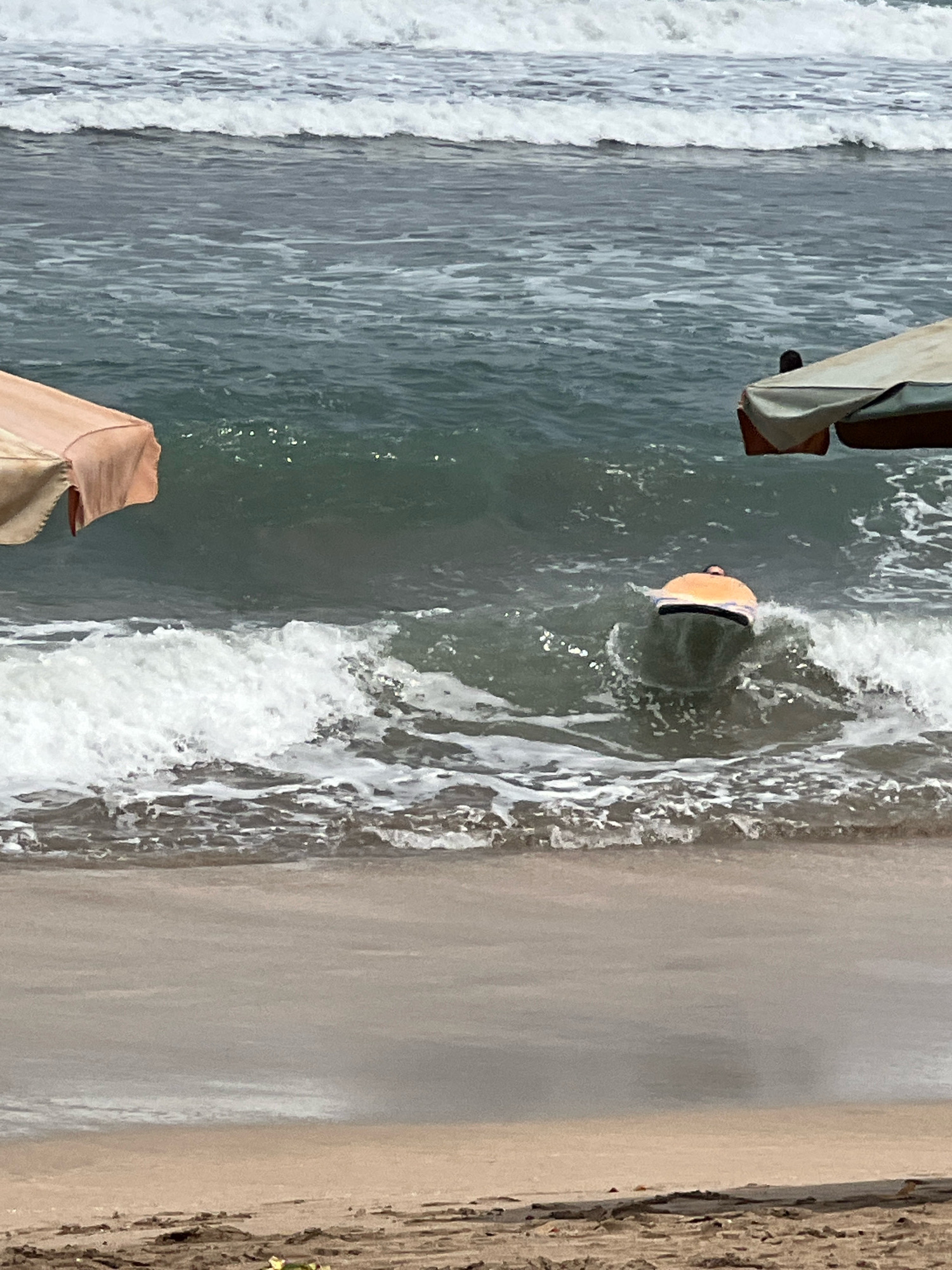 The image depicts a beach scene in Kuta, Indonesia. The ocean is visible with waves breaking on the shore. In the water, a person is lying on a boogie board, facing away from the camera, and riding a wave. Two beach umbrellas are partially visible at the top corners of the frame, suggesting a public beach setting. The sand in the foreground is light brown and appears to have some debris. The overall lighting suggests it is daytime, possibly overcast.