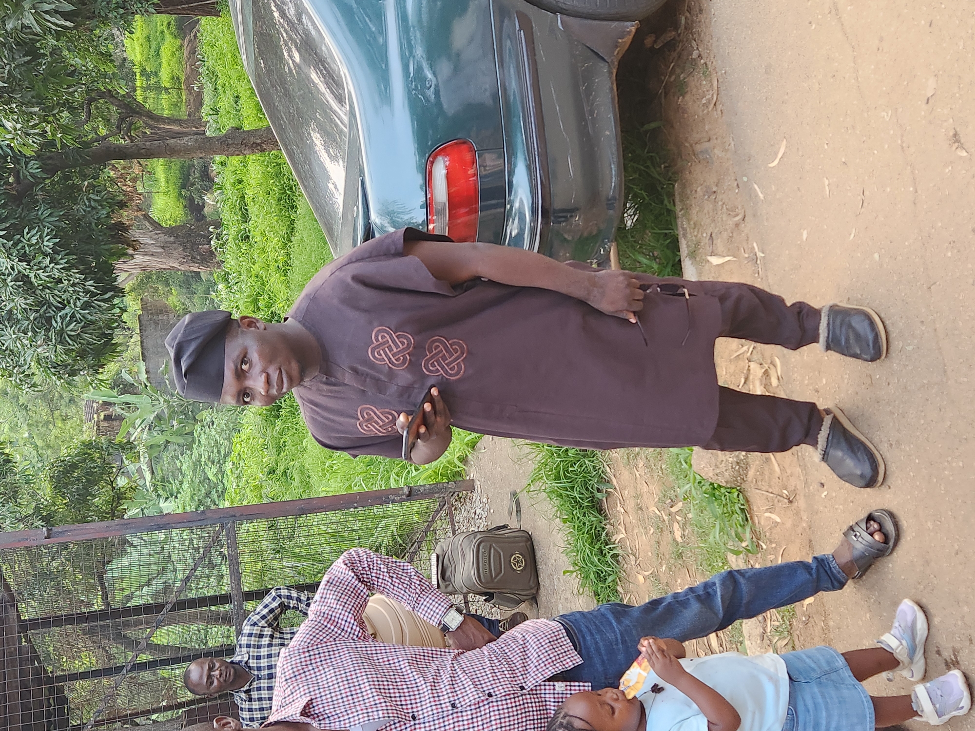 In Jos, Nigeria, an outdoor scene depicts three individuals. A man in the foreground, dressed in a dark brown traditional garment and a black cap, holds a smartphone and a pair of eyeglasses. To his left, another man wears a red and white plaid shirt and blue jeans. Partially visible to the left is a young child in a light blue t-shirt and denim skirt, eating a snack. Behind them, a man in a checkered shirt stands near a metal fence. A dark green car is parked to the right, and a backpack rests on the ground near the man in the plaid shirt. The environment features lush green vegetation and a dirt path.