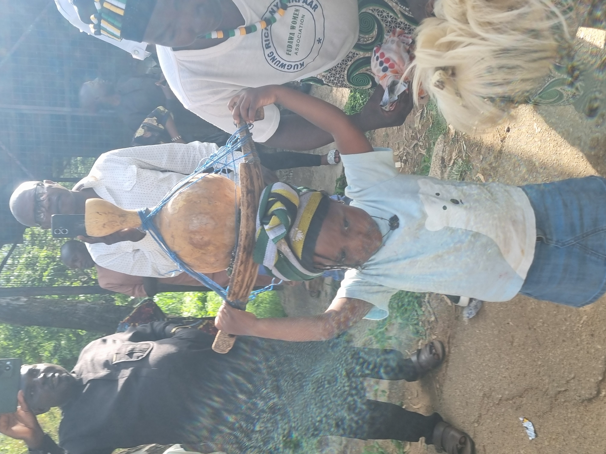 The image depicts an outdoor gathering, likely in Jos, Nigeria. Several people are present, with a focus on a young child in the foreground holding a gourd attached to a wooden stick, which is further secured by blue netting. The child is wearing a striped hat and a light blue t-shirt. To the left, an adult in a white shirt is visible, holding a dark-colored phone. Another adult in a black shirt is also holding a phone. In the background, others are partially visible, including someone wearing a white t-shirt with text on it. The setting appears to be natural, with greenery and a dirt or gravel path. The lighting suggests it is daytime, possibly late morning or early afternoon. The overall mood seems to be communal, with people engaged in an activity or event. Visible text on the white t-shirt reads "AFRICA NY IRKPAAR FUDUWA WOMEN ASSOCIATION KUGWUNG".