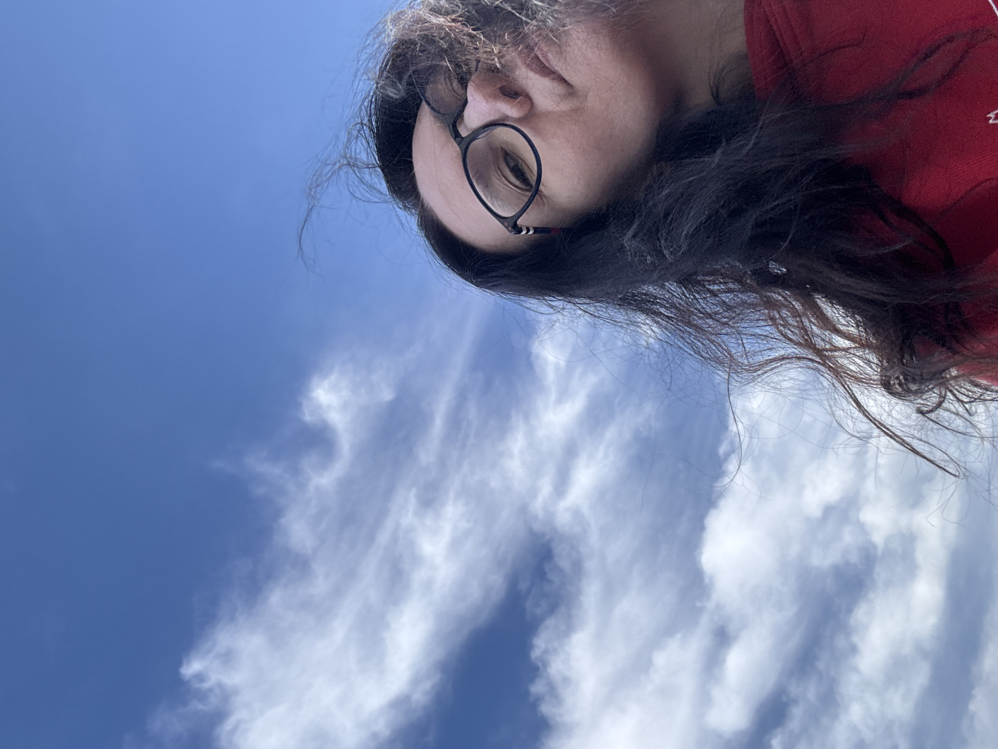 A low-angle shot shows a person wearing glasses and a red garment, with dark hair blowing across their face. The background consists of a bright blue sky scattered with wispy, white clouds. The location cannot be determined from the image.