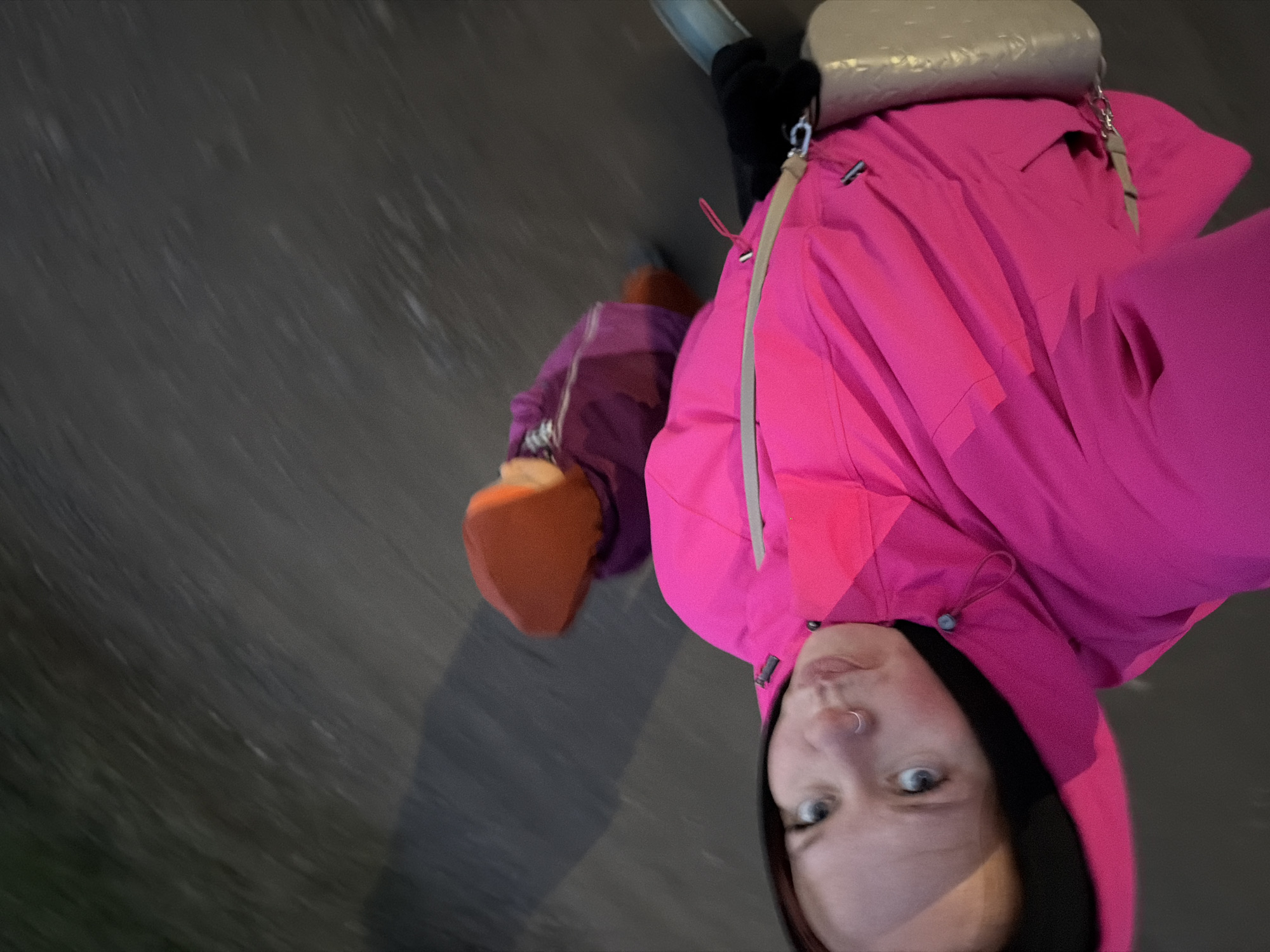 The image, captured from a tilted, high-angle perspective, features a young adult, likely female, looking directly into the camera. She has fair skin, light-colored eyes, and a nose ring. She is dressed in a vibrant fuchsia pink hooded jacket with grey accents and drawstrings. A light beige shoulder bag with a metallic chain strap hangs over her right shoulder (viewer's left). A black glove or sleeve is partially visible on her left hand.
The background is a dark, blurry, textured surface, possibly a wet road or pavement, with visible horizontal streaks that suggest motion blur or light precipitation like rain or snow. This low-light setting indicates the photo was taken during the evening, night, or on a heavily overcast day in Chaikovsky, Russia. A partial view of another individual is seen in the lower left, identified by a purple garment and an orange shoe or glove, implying close proximity to the main subject.
The subject's expression is neutral and direct. The image conveys a sense of movement within a cool or cold outdoor environment, consistent with the protective outerwear. There is no visible text.