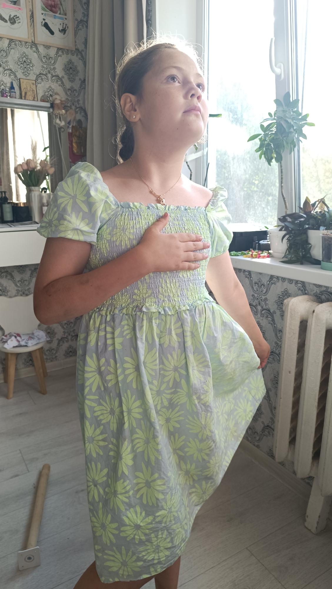 A young girl with light brown hair, wearing a pale green and light blue floral dress, stands in front of a window. Her right hand is placed over her chest, and she is looking upwards and to the right. Sunlight streams in from the window, creating a bright glow around her head and shoulders. Behind her, on the windowsill, are several potted plants and some small decorative items. To the left of the girl, a mirror reflects a shelf with items. The flooring is light-colored wood. A white radiator is visible on the right side of the image. This scene appears to be indoors, possibly in a residential setting in the city of Chaikovsky, Russia.
