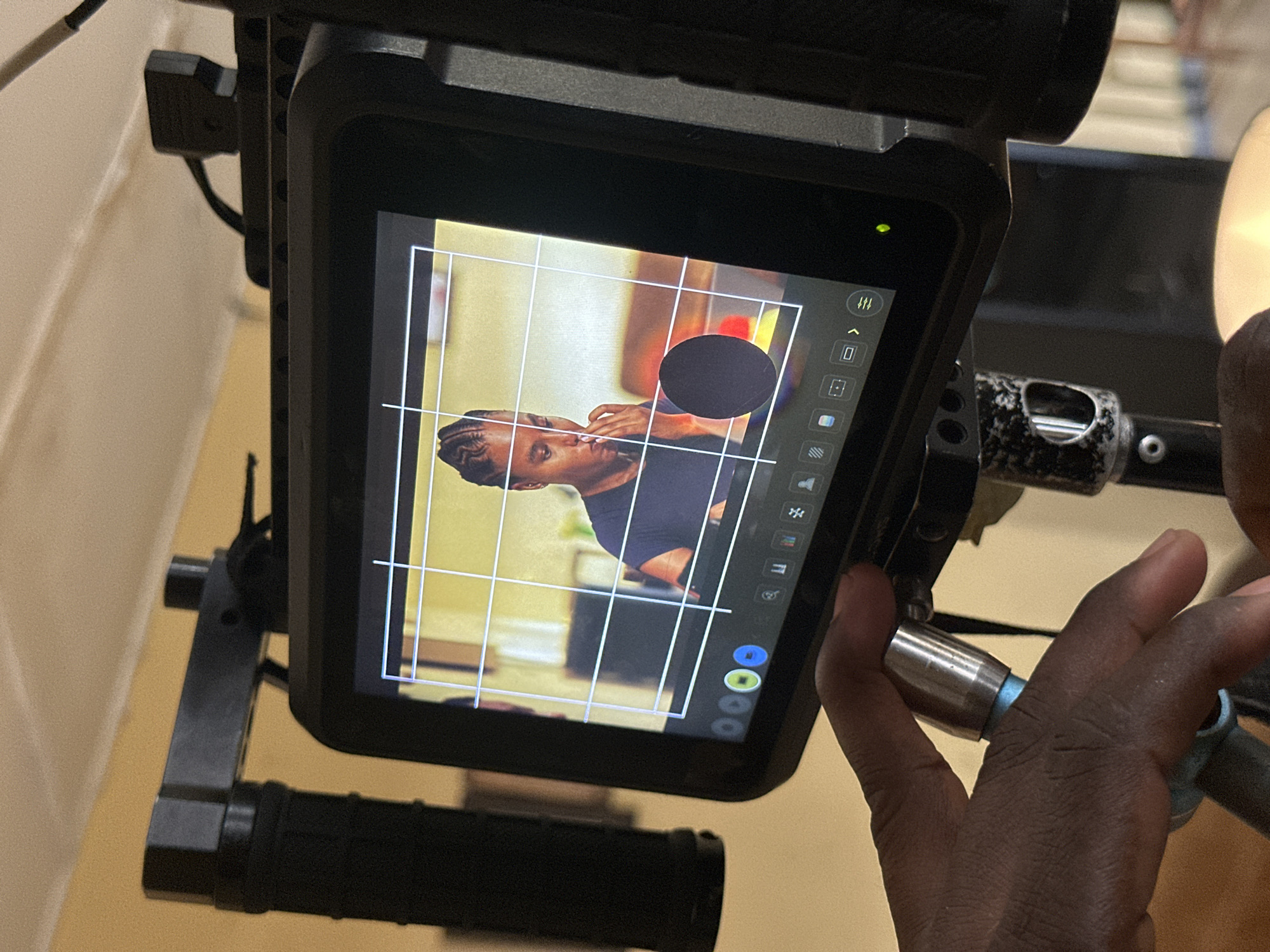 The image displays a close-up of a digital camera monitor, likely an external field monitor, mounted on what appears to be a camera rig. The monitor's screen shows a live feed or recorded image of a person, identifiable by their dark skin, braided hair (cornrows), and dark t-shirt. The person is captured in a profile view, looking left, with one hand near their face. A white grid, likely a compositional guide, is overlaid on the image. A solid black circular shape is superimposed over the display, partially obscuring the background behind the subject.
Along the right edge of the monitor's screen, a column of icons indicates various camera or monitor functions, including symbols for settings (a wrench), framing or aspect ratio, and three colored circles (green, blue, yellow) at the bottom. A small green indicator light is visible on the upper left corner of the monitor's black bezel.
A dark-skinned hand is visible on the right, gripping a metallic part of the camera rig, suggesting human interaction with the equipment. The monitor is housed within a sturdy black frame, integrated into other camera accessories like handles. The actual background behind the monitor is blurred, indicating an indoor setting with light-colored walls. This scene depicts a technical setup for video production or filmmaking, with the provided context placing the activity in Lagos, Nigeria.