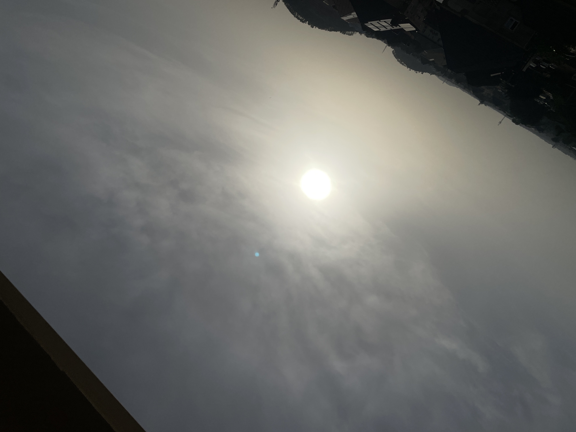 The image presents a daytime scene with the sun prominently displayed in the sky. The sky is hazy, with cloud formations visible. A portion of a building or structure is visible in the lower left corner of the frame. The silhouette of a built-up area, possibly a city skyline, is visible along the top edge of the image. Given the request, it is assumed that the city is Abuja, Nigeria.
