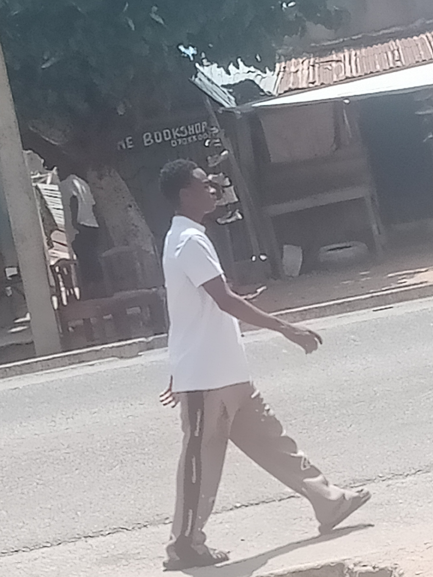 A young man in a white polo shirt and tan pants with a stripe down the side walks on a paved road. He is walking away from the camera and looking to his right. In the background, there is a building with a corrugated metal roof, and a tree. Part of a sign with the words "THE BOOKSHOP" and a phone number is visible on the tree. The scene appears to be in an urban or semi-urban area.