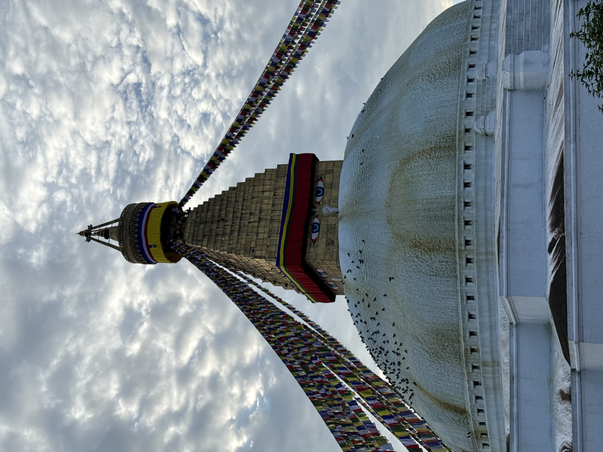 A low-angle, medium shot captures the Boudhanath Stupa in Kathmandu, Nepal, under a cloudy sky. The stupa's large, white dome dominates the lower half of the frame, its surface textured and stained in places. Numerous small, dark birds are perched on the dome. A band of small square windows runs around the base of the dome. Above the dome, a golden, multi-tiered spire rises, adorned with colorful prayer flags strung across its width. The spire features large, blue eyes painted onto its surface, a common motif on Buddhist stupas. The sky is filled with a mix of white and grey clouds, with patches of lighter sky visible. The scene is devoid of any people.