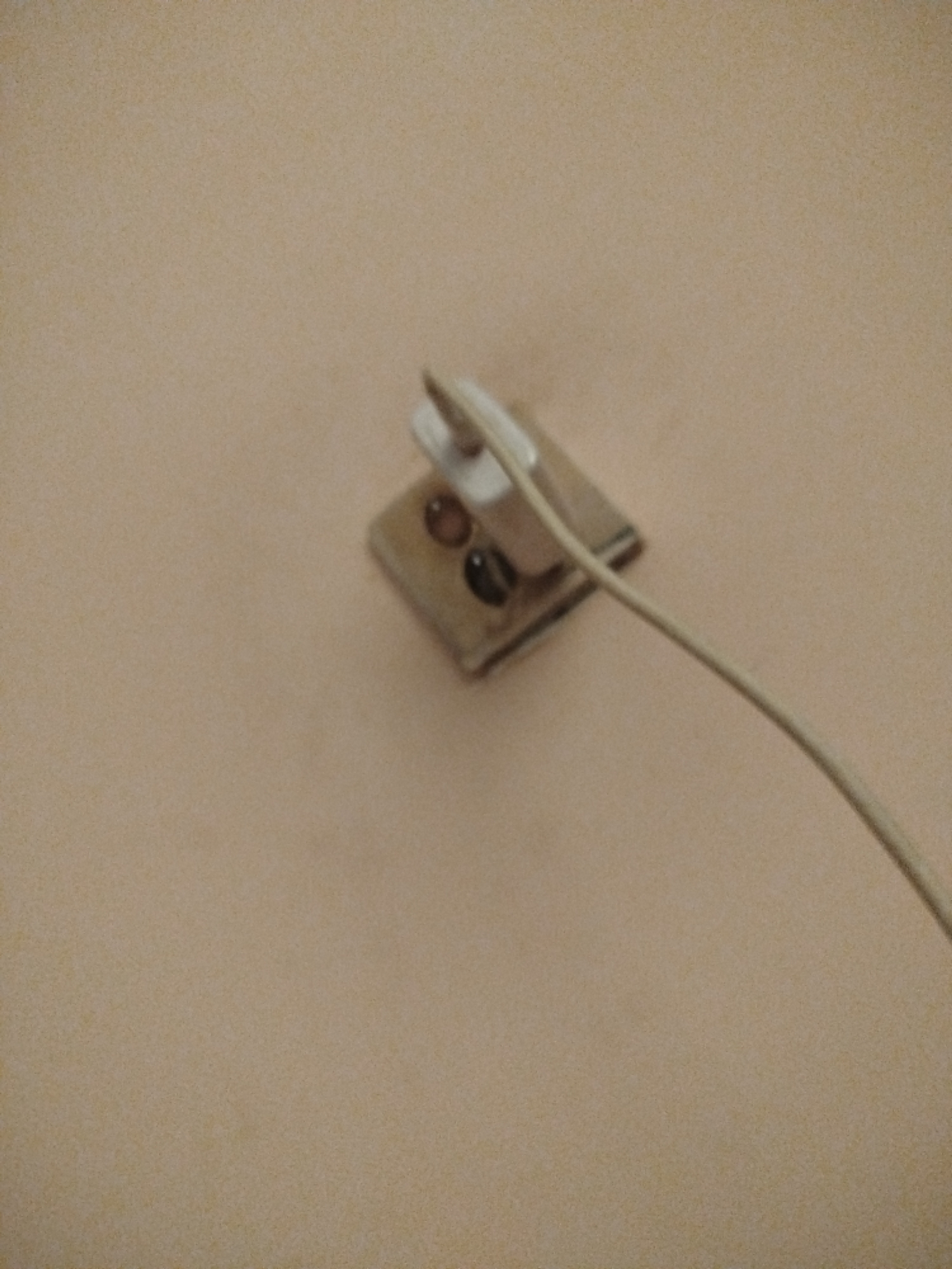 An electrical plug is inserted into a wall socket. The plug is white and has two prongs. The wall socket is square and beige, with two circular openings. A thin, light-colored wire extends from the plug and curves across the frame. The background is a plain, light brown wall. There are no people or other objects visible in the image. The location is not specified.