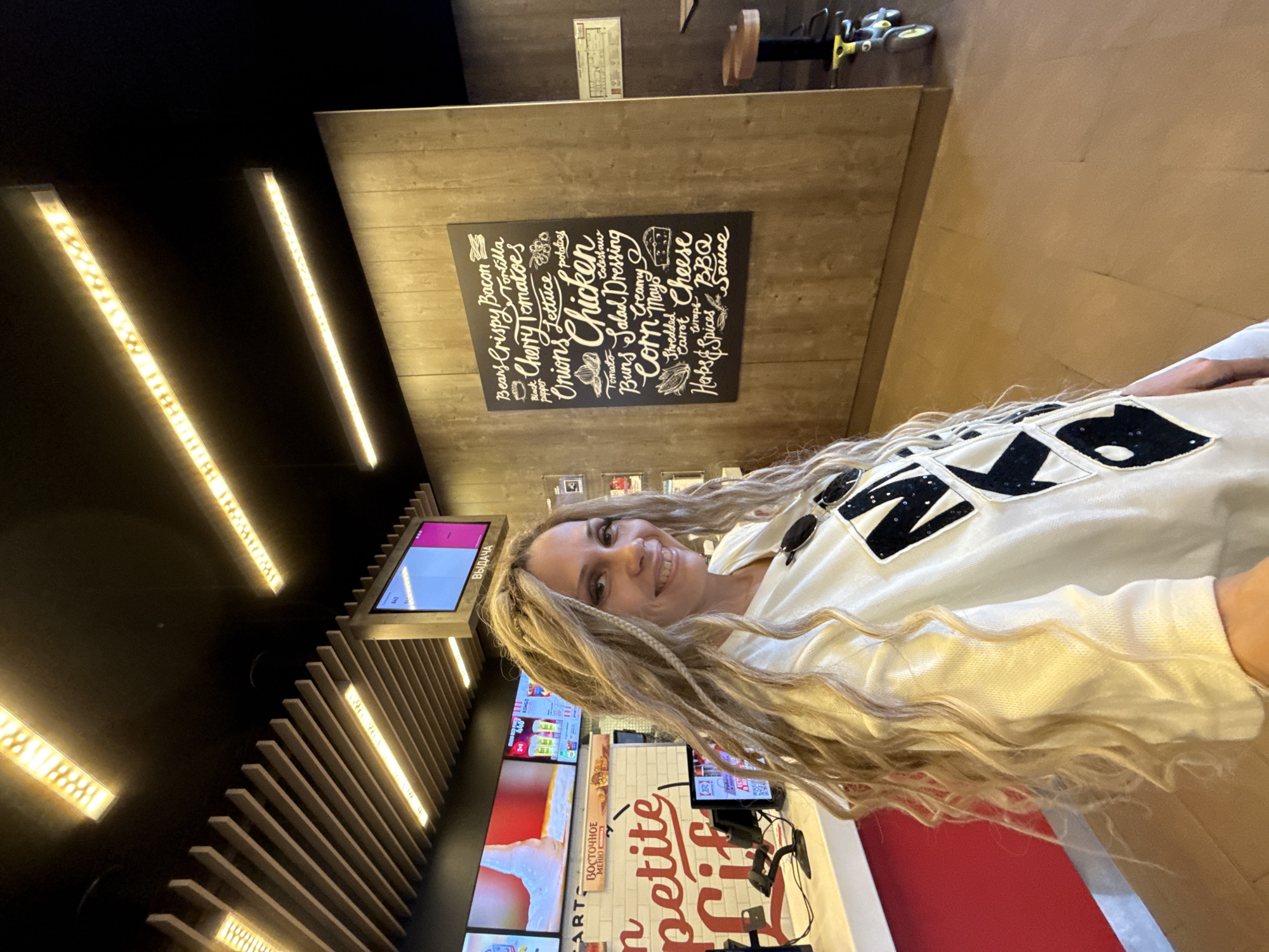 The image depicts an indoor scene within a modern fast-food or casual dining establishment, located in Rusanovo, Russia. A fair-skinned adult female is prominently featured in the foreground, facing slightly left and smiling directly at the viewer. She has long, wavy blonde hair interwoven with several thin braids or dreadlocks, cascading over her shoulders and chest. Her eyes are accentuated with dark eyeliner. She is wearing a white long-sleeved top or sweatshirt adorned with large black, irregularly shaped patches, some of which appear to be sequined, on the front. A pair of black sunglasses is hooked onto the neckline of her top. Her right arm is partially visible on the bottom right, suggesting she is holding the camera or phone for a selfie.
The background on the left reveals a service counter with a red base and a white top. Above the counter, several large flat-screen monitors display digital menus and promotions, including images of beverages and food items. One screen above the main counter shows "ВОСТОЧНОЕ МЕНЮ" (Eastern Menu) in red text above a stylized "Bon Apetite Life" logo. Further right, another screen displays "ВЫДАЧА" (Issuance/Pickup) in Cyrillic. On the counter, various point-of-sale equipment, including card readers and a screen displaying a QR code, are visible.
The background on the right features a wall clad in vertical wooden paneling. Mounted on this wall is a large, rectangular black chalkboard-style menu board with white handwritten-style text listing ingredients and dishes such as "Crispy Bacon," "Cherry Tomatoes," "Onions," "Chicken," "Salad Dressing," "Corn," "Cheese," and "BBQ Sauce." Beyond the wooden wall, a glimpse into another section of the establishment reveals a dark space with a light brown bar stool and a child's yellow and gray tricycle-like toy on the floor.
The ceiling above is dark black, with numerous long, rectangular LED light fixtures providing bright, warm illumination. Some lights are positioned individually, while others are integrated into a slatted wooden ceiling structure that runs diagonally across the overhead space. The floor is covered with light brown or tan tiles. The overall environment appears clean, modern, and well-lit.