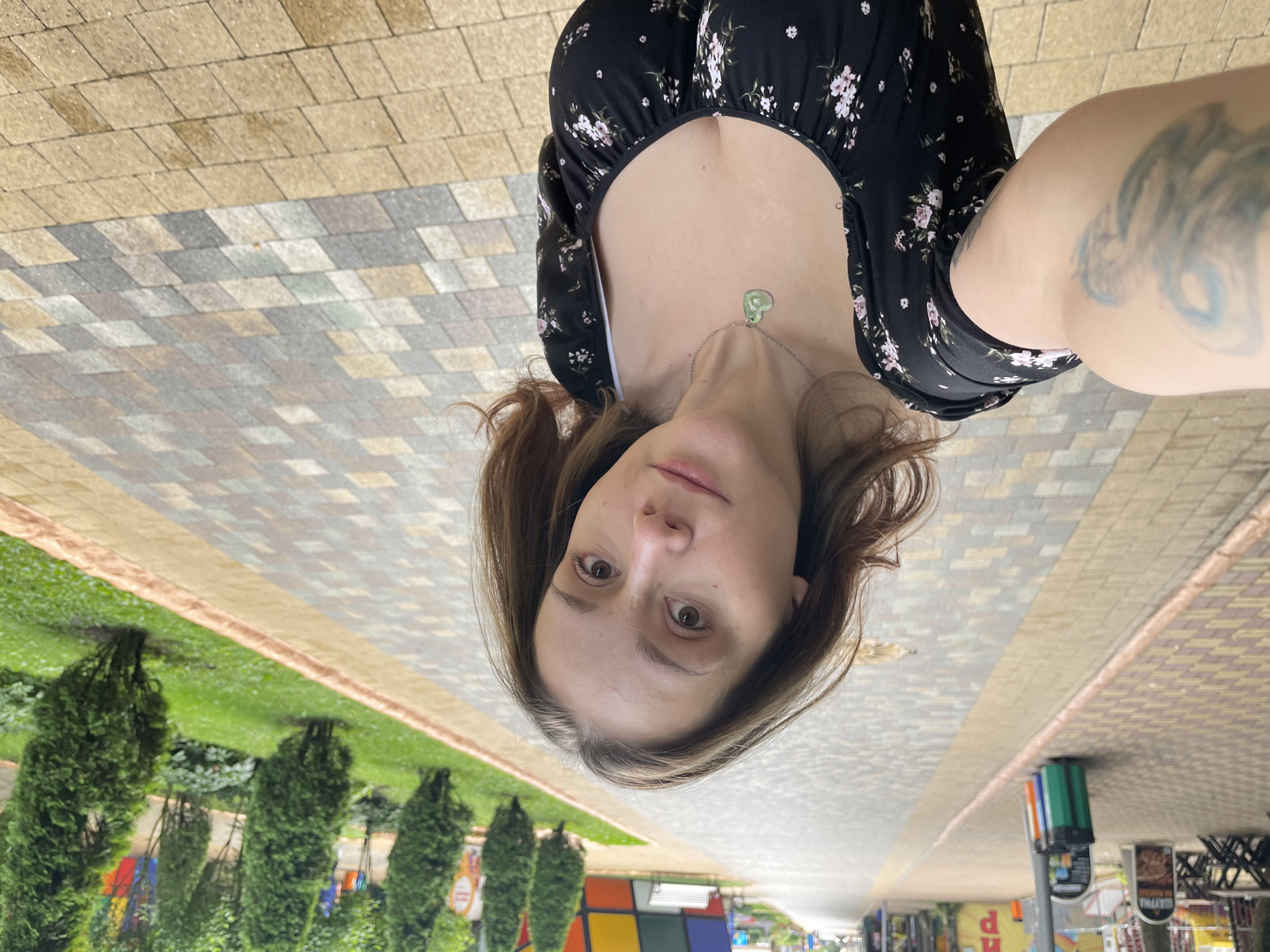 A woman with brown hair, wearing a black floral dress and a green heart-shaped necklace, takes a selfie in an outdoor setting. She has a tattoo on her left arm, which is visible on the left side of the frame. The background shows a paved walkway with a checkered pattern, flanked by a grassy area with manicured cypress trees and other greenery. In the distance, there are buildings with colorful facades, benches, and trash receptacles, suggesting a park or public area. The overall scene is bright and appears to be during daytime. The location is identified as the "Park Pobedy" (Victory Park) in Stavropol, Russia, based on visual cues including the architectural style of the distant buildings and the type of greenery commonly found in Russian parks.