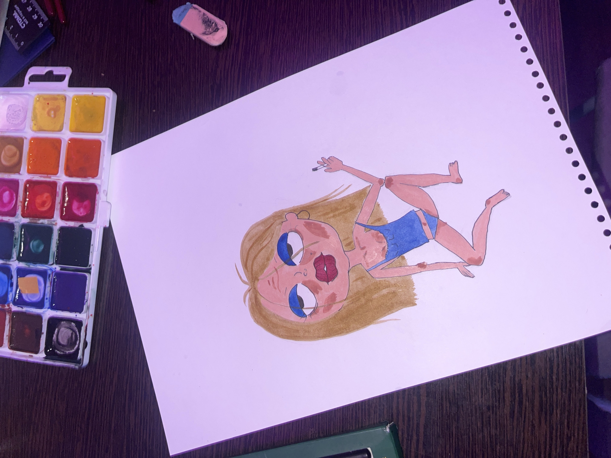 The image shows a drawing of a woman in a blue swimsuit, lying on her back on a white sheet of paper. The drawing is in a cartoonish style and appears to be colored with watercolors. The woman has blonde hair, blue eyes, and large lips. She is holding a cigarette in her right hand. Next to the paper is a watercolor palette with various colors, a ruler, and an eraser. The scene is on a brown wooden table. There are no indications that this image is in Saint Petersburg, Russia.